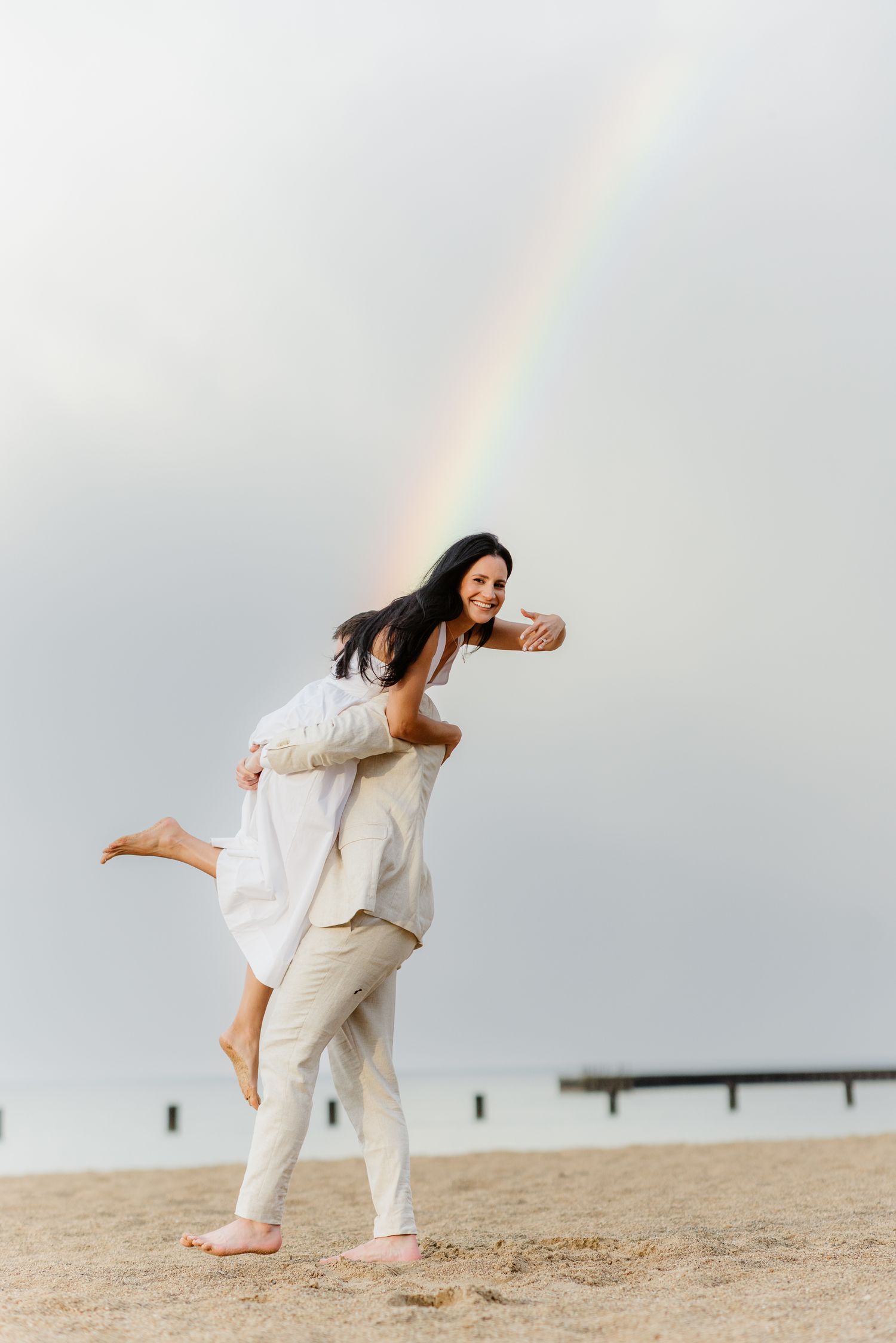 A laughing woman gets a piggyback ride from her partner on a beach with a rainbow in the background.