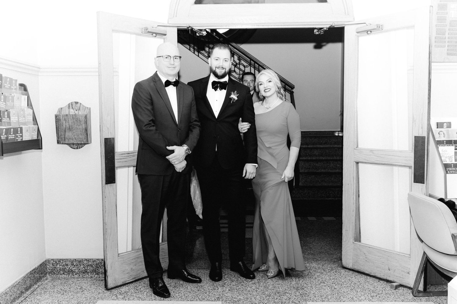 Black and white portrait of groom with parents at wedding venue entrance doorway.