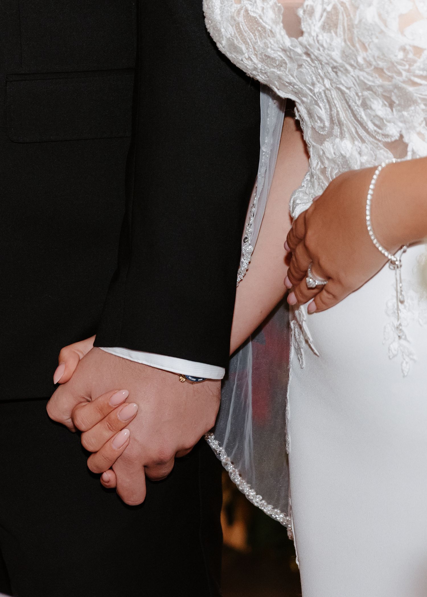 Close-up of bride and groom holding hands showing wedding ring, pearl bracelet, and lace wedding dress detail.