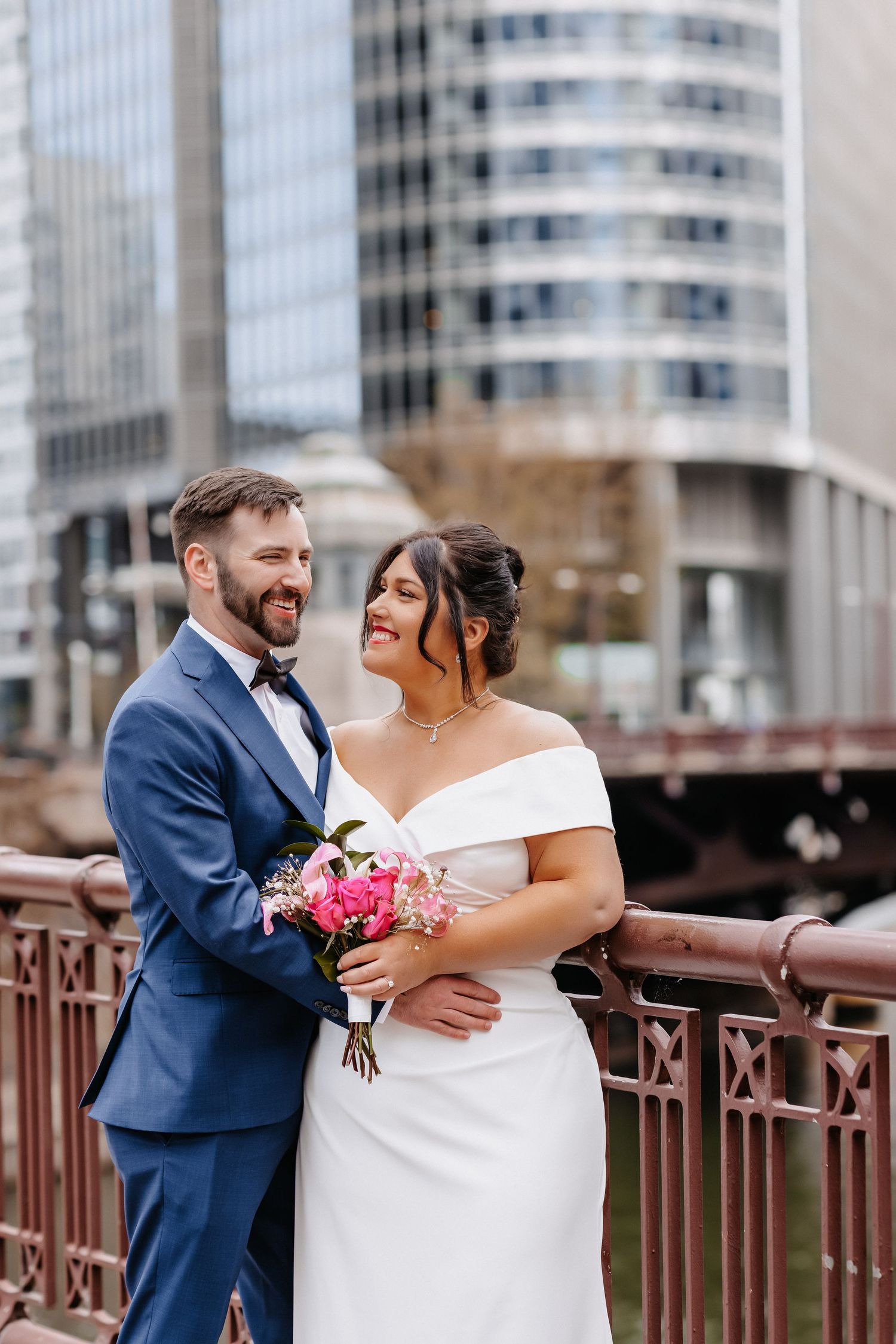 Bride and groom share romantic moment on city bridge, she holds pink bouquet, skyscrapers behind.