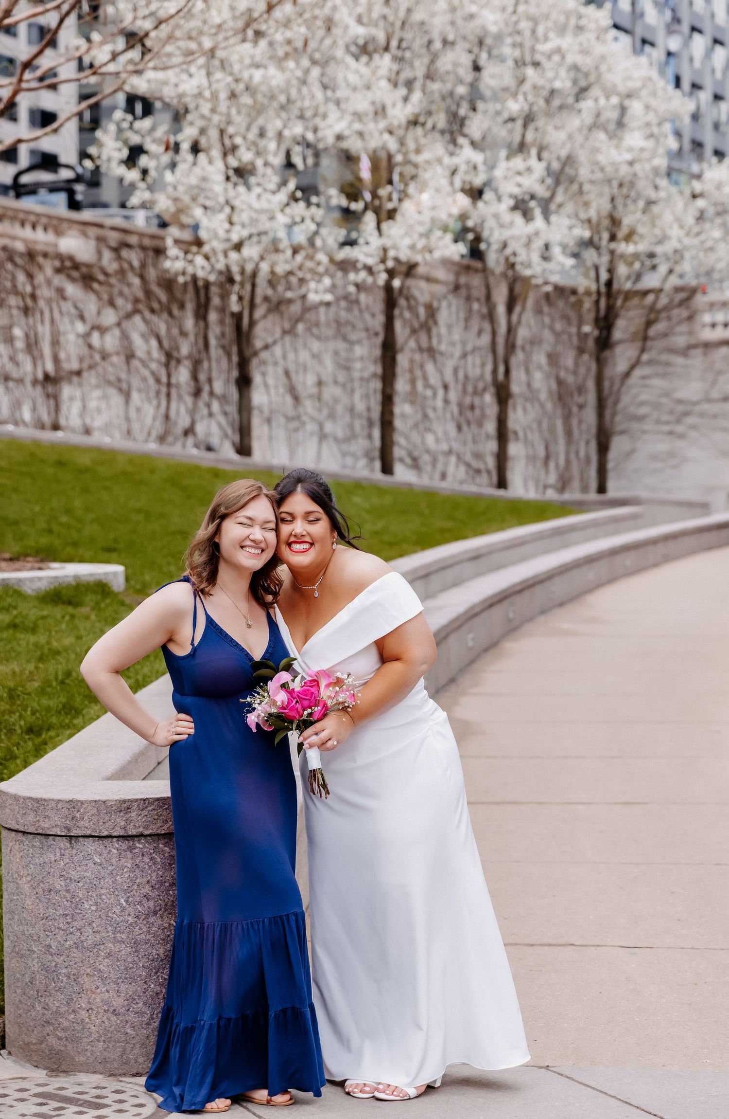 Bride in white off-shoulder dress and bridesmaid in navy gown smile together near flowering trees.