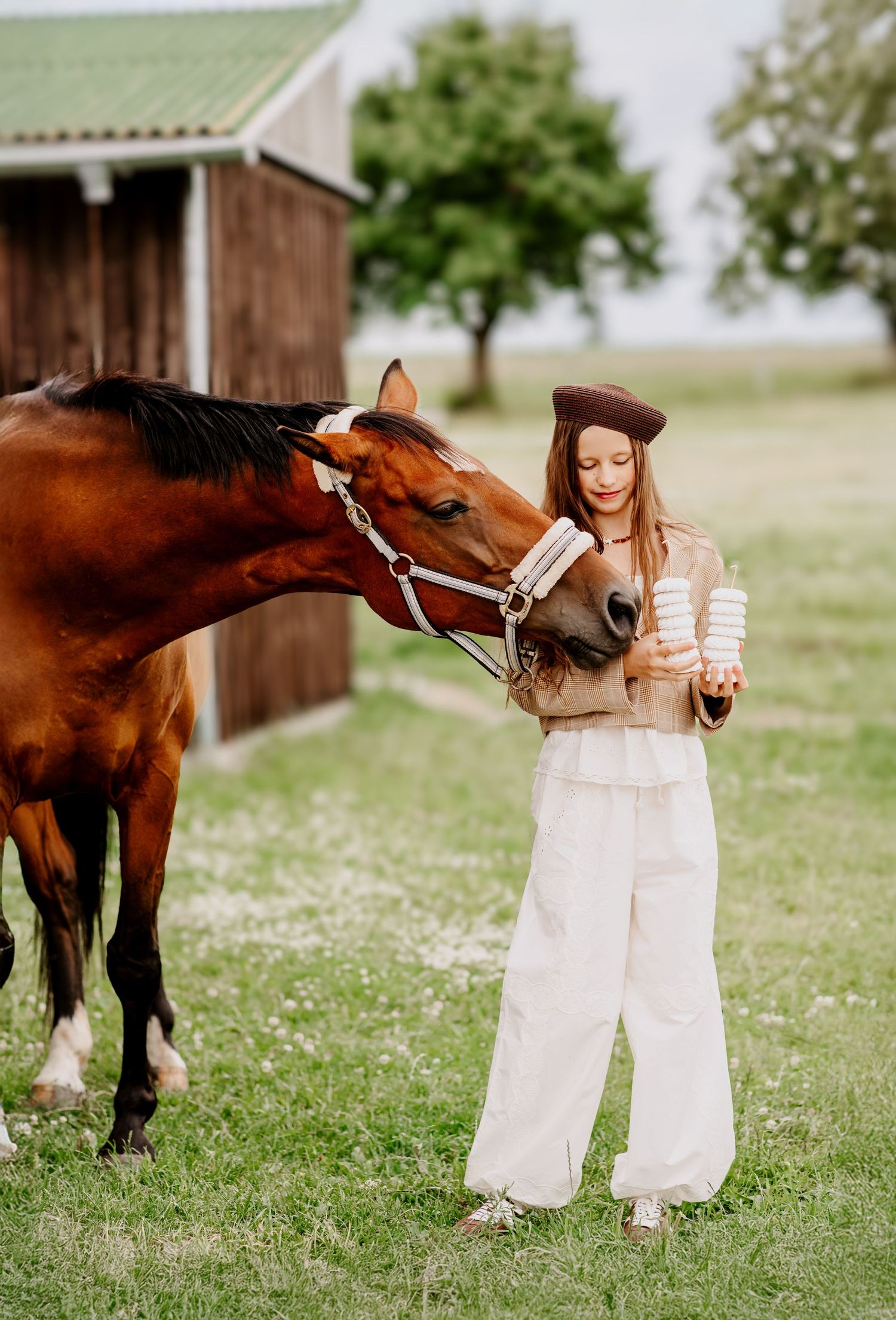 Smiling young woman in beret feeds a chestnut horse from her hand near a wooden barn.