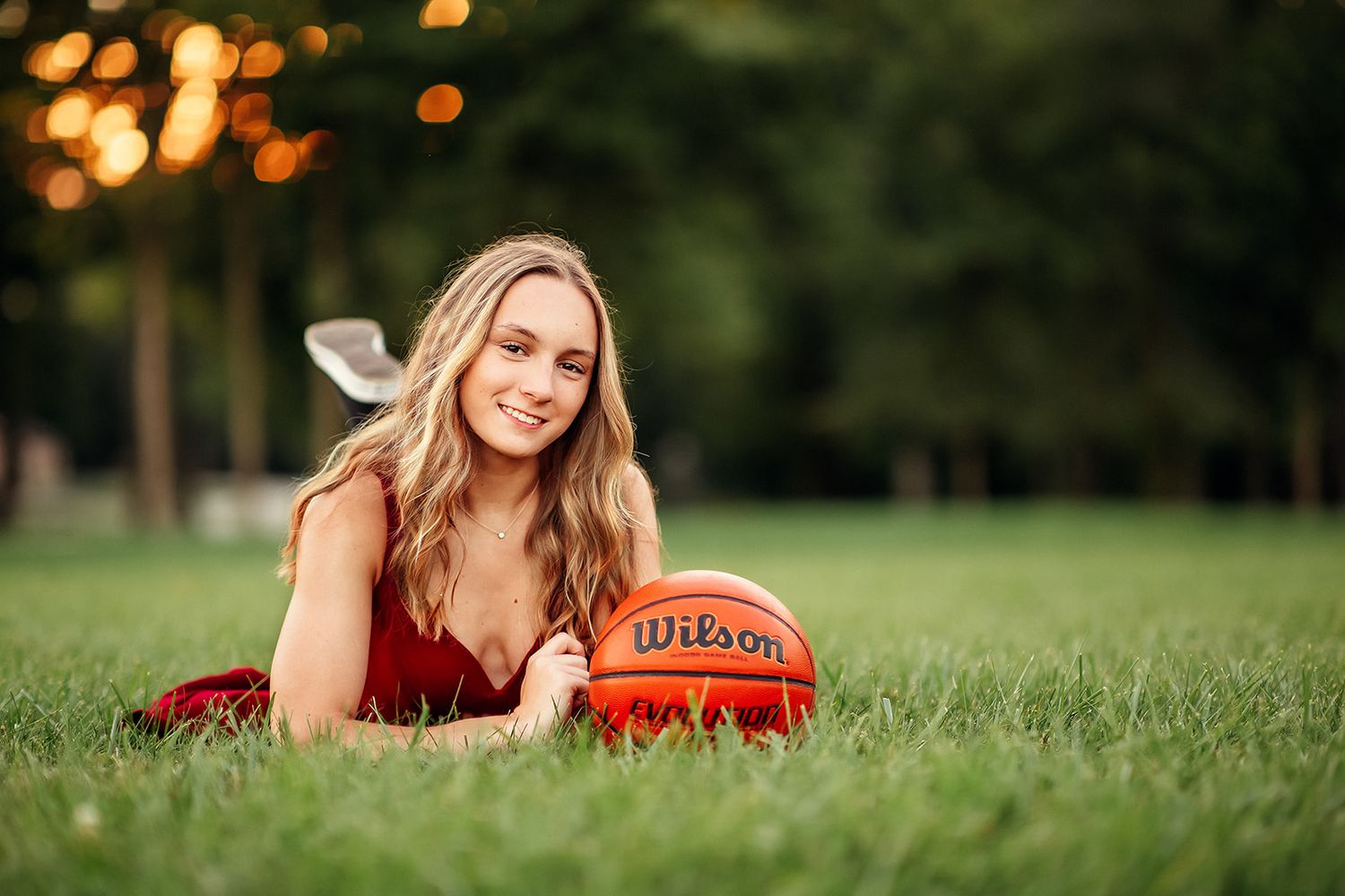 Smiling blonde girl in red dress lying on grass with a Wilson basketball.