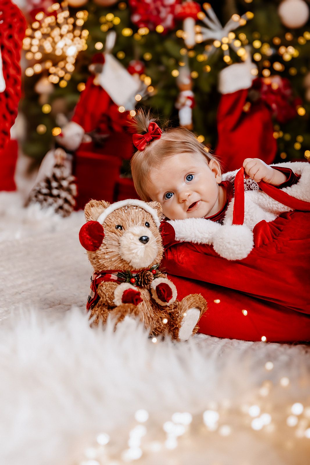 Baby girl in Santa outfit lying with a festive teddy bear near a decorated Christmas tree with bokeh lights.