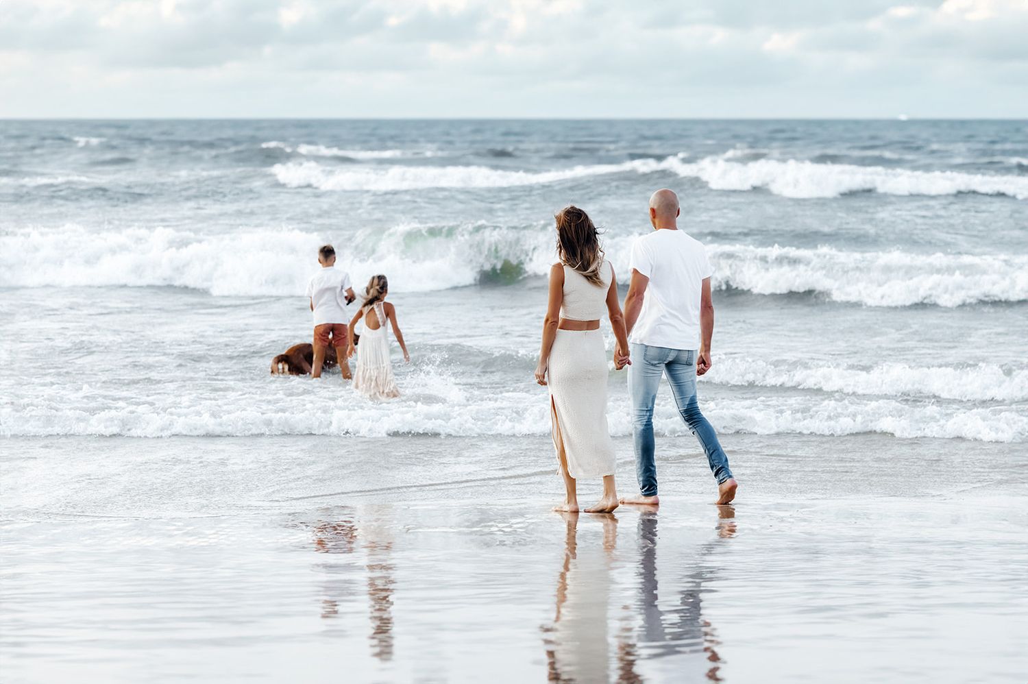 A couple walks hand-in-hand on a scenic beach while children play in the waves in the background.
