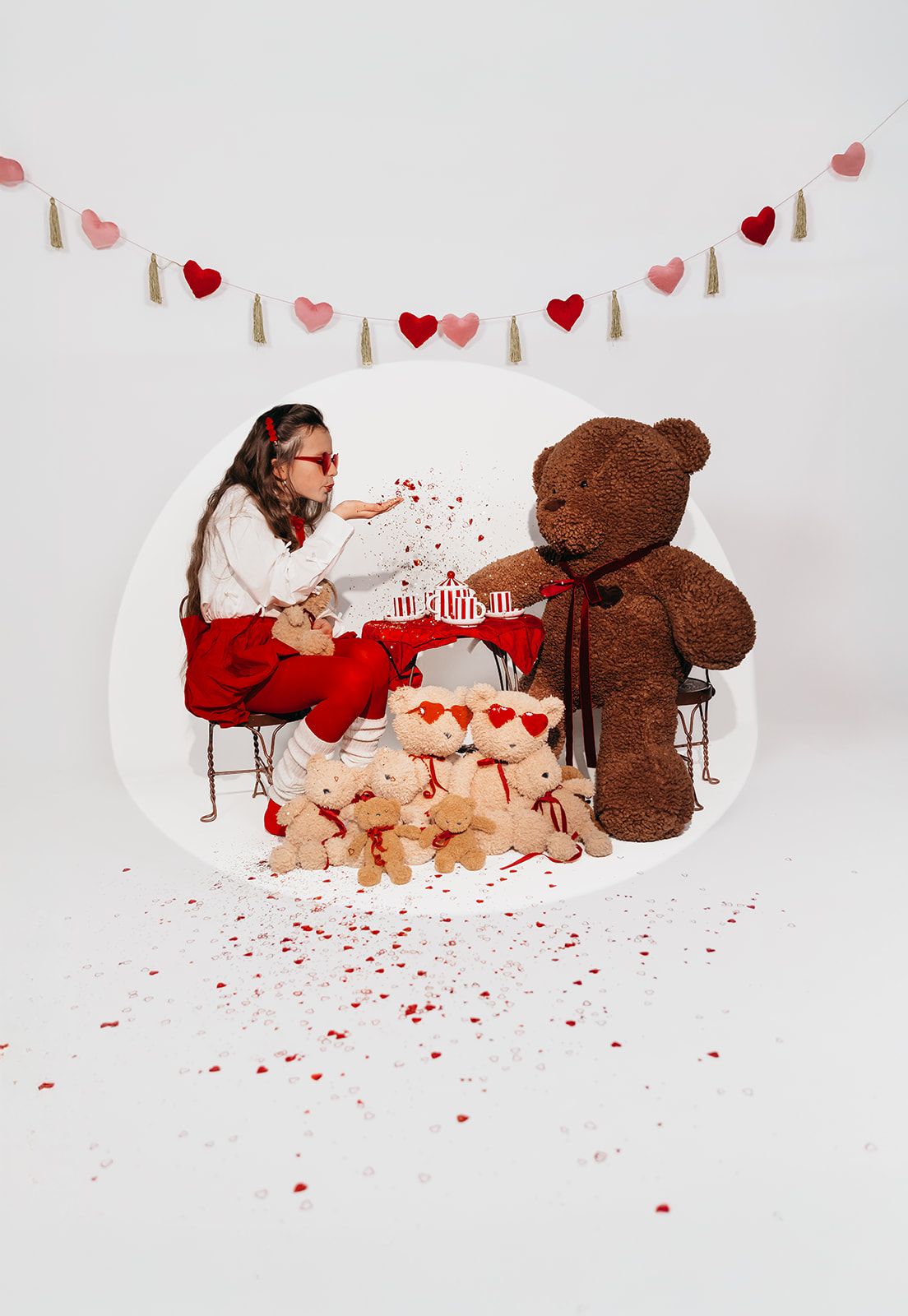 Girl blowing kisses at giant teddy bear surrounded by small bears, heart garland, and confetti in Valentine's Day photo.