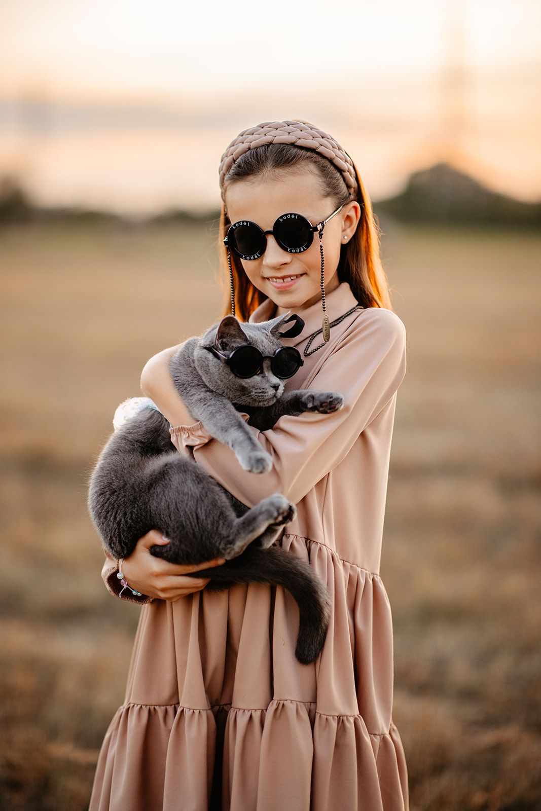 Smiling girl in beige dress holding gray cat wearing sunglasses in golden field at sunset.