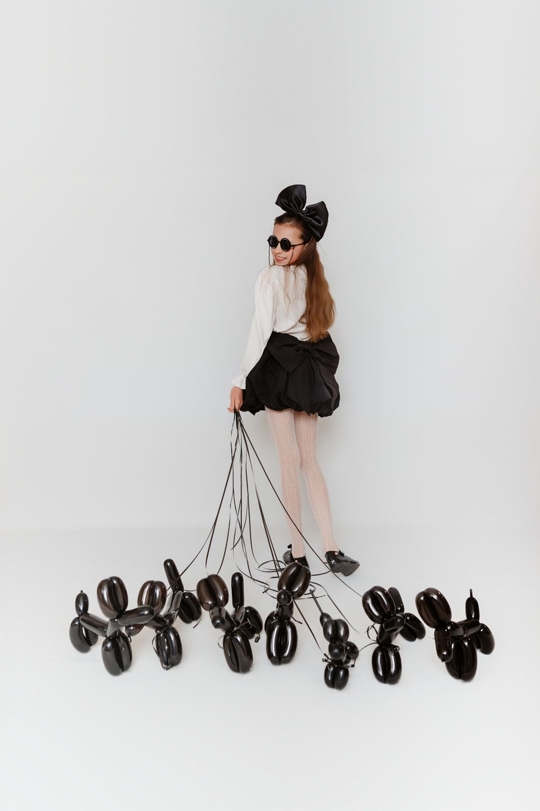 Young girl in black skirt and sunglasses holds strings of black balloon dogs on white background.