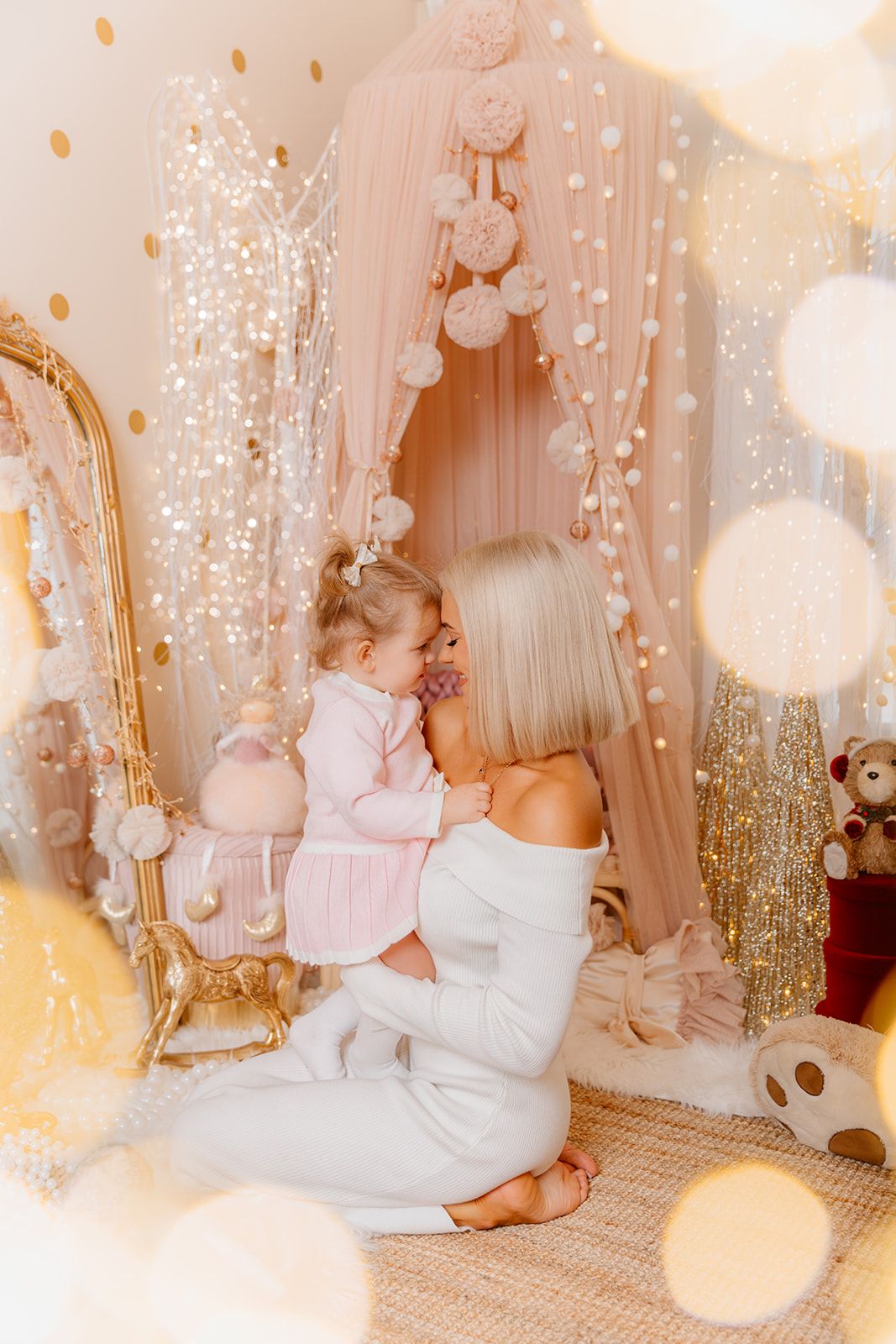 A blonde mother holds her toddler daughter in a dreamy pink nursery decorated with fairy lights and pompoms.