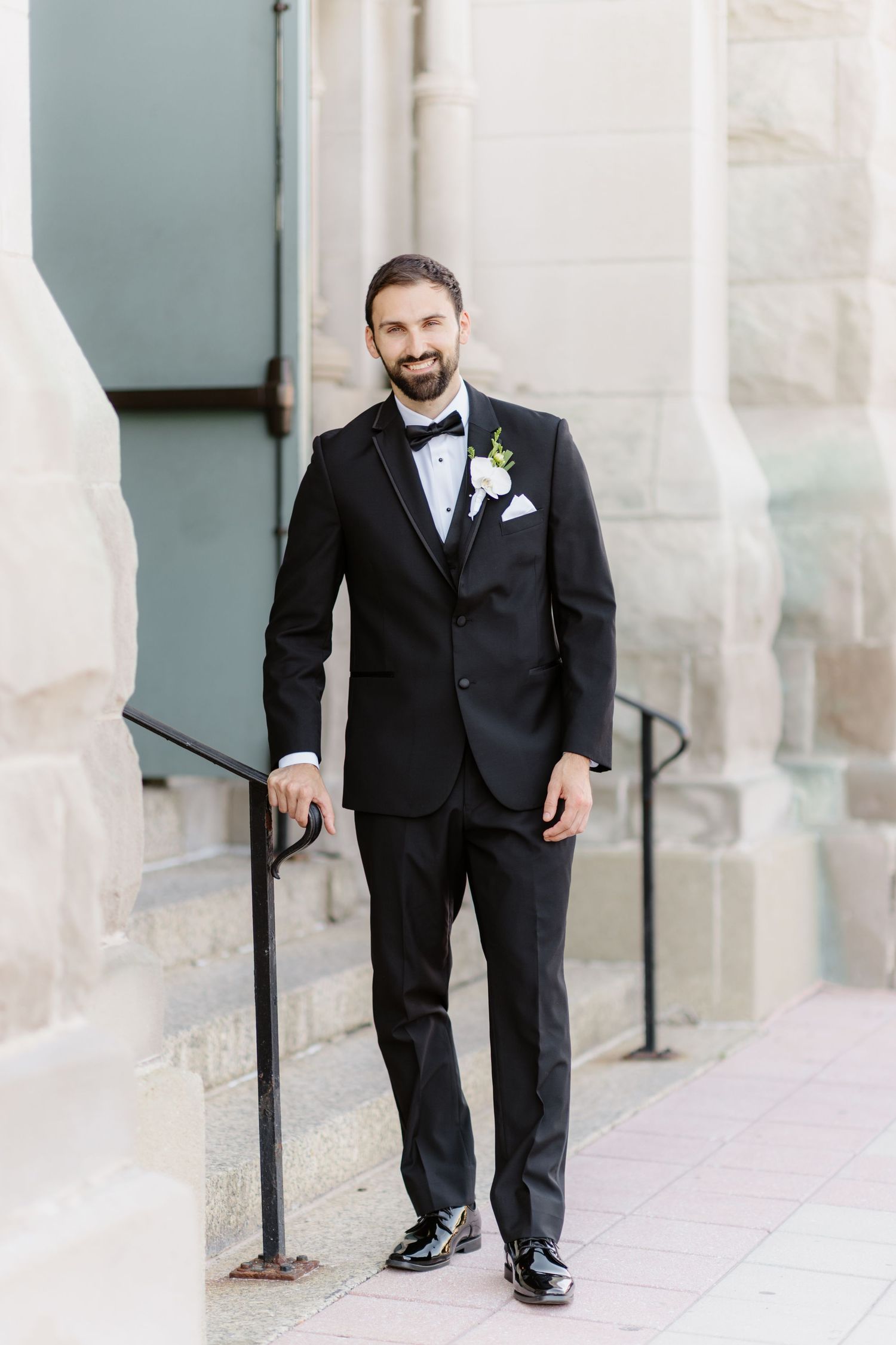 Groom in black tuxedo with white boutonniere smiling outside stone building.