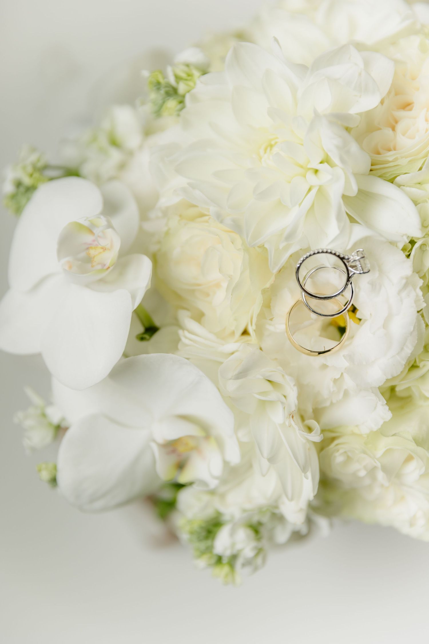 Two gold wedding bands resting on a lush white bridal bouquet of peonies and orchids.
