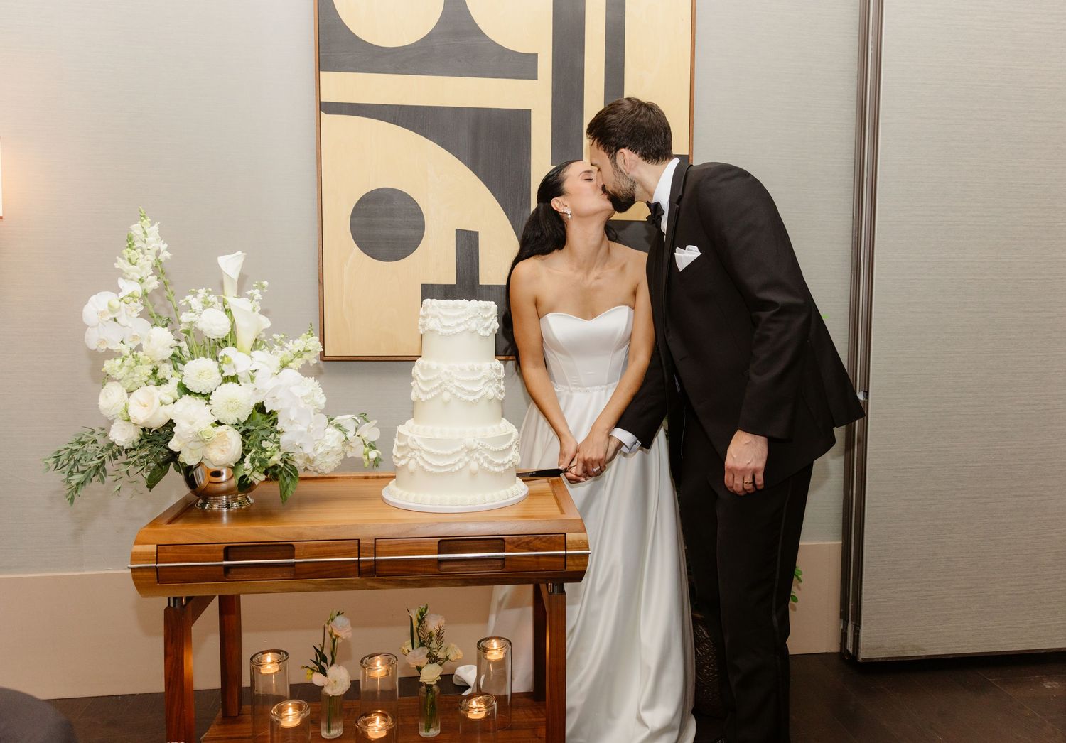 Bride and groom sharing a tender kiss beside their white tiered wedding cake at indoor reception.