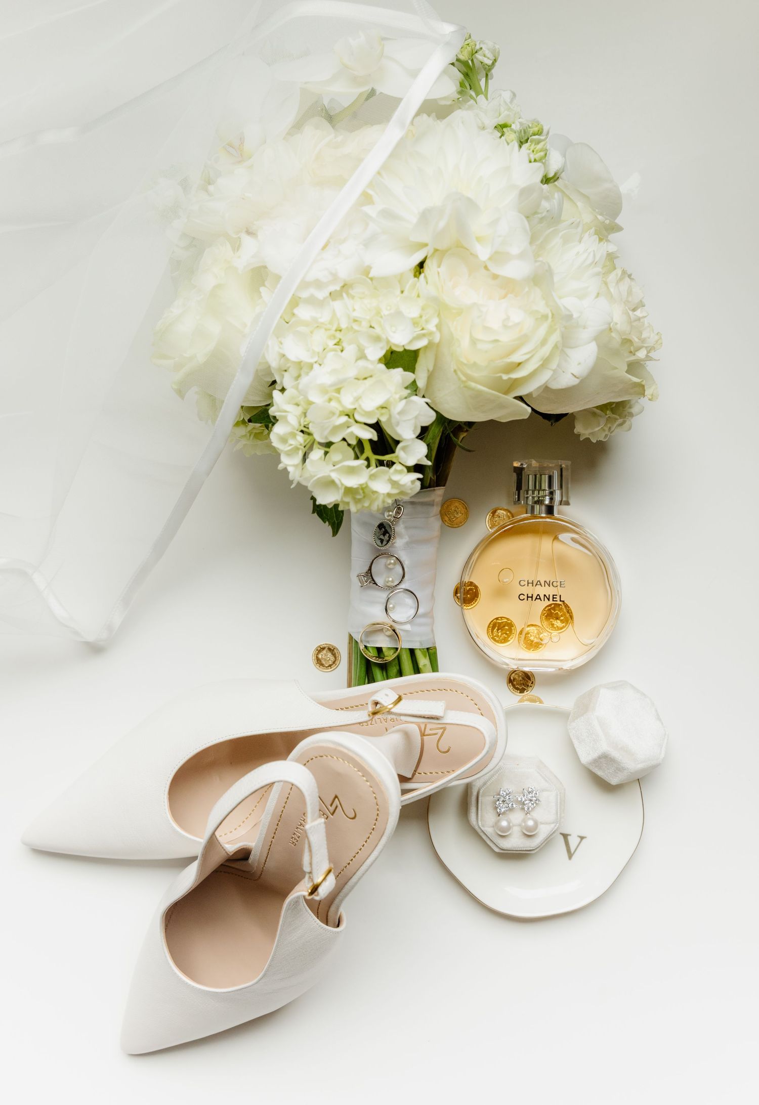 Bridal flat lay featuring white heels, Chanel perfume, white floral bouquet, veil, and ring box.