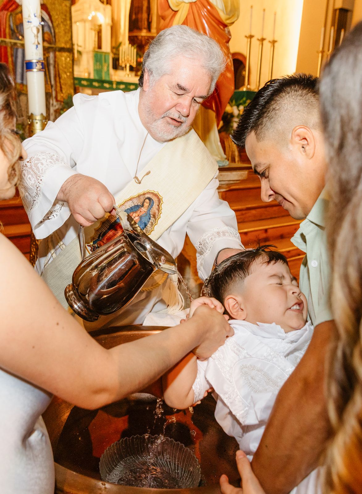 Catholic priest performing baptism rite pouring water over infant held by father in ornate church.