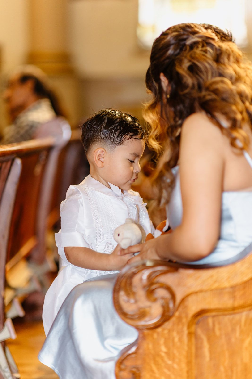 Young boy in white outfit holds stuffed animal while sitting in church pew during ceremony.
