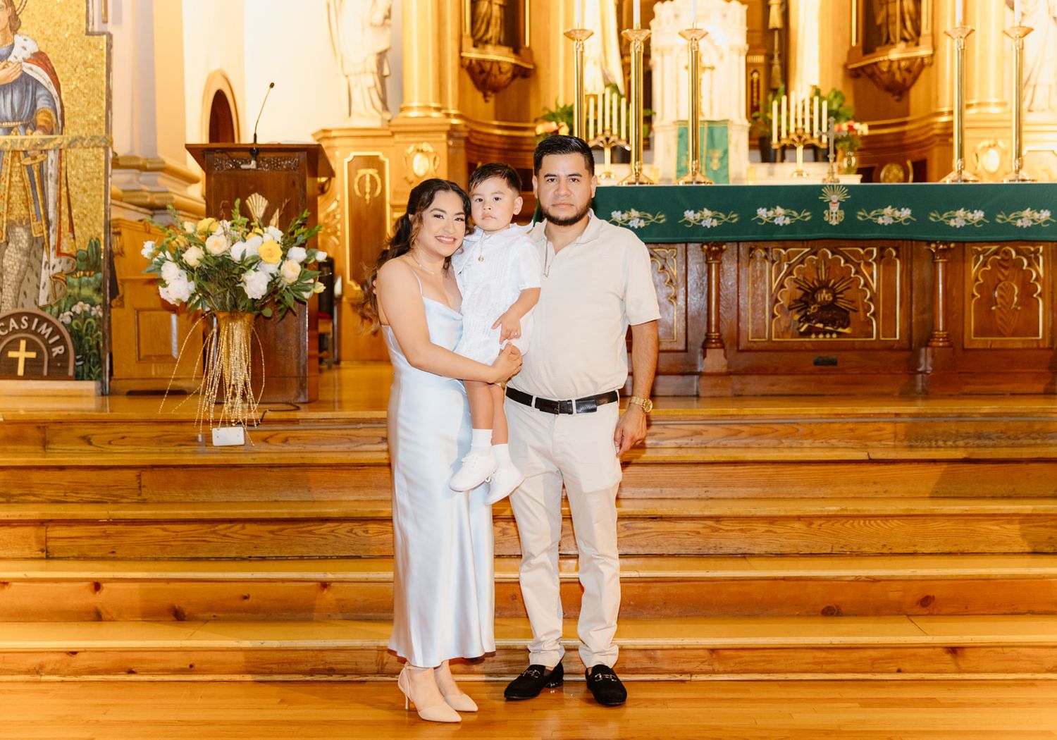 Family of three posing in a beautiful wooden church during a baptism ceremony, mother in white dress holding baby.