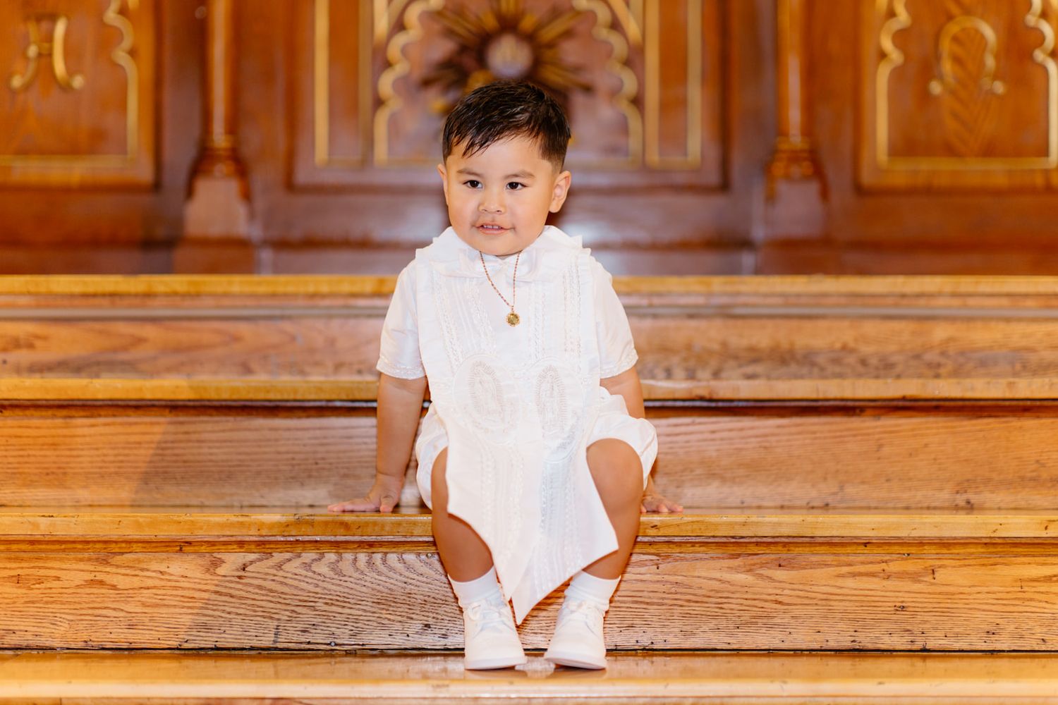 Toddler boy in white barong tagalog sits on ornate wooden church steps wearing a gold necklace.