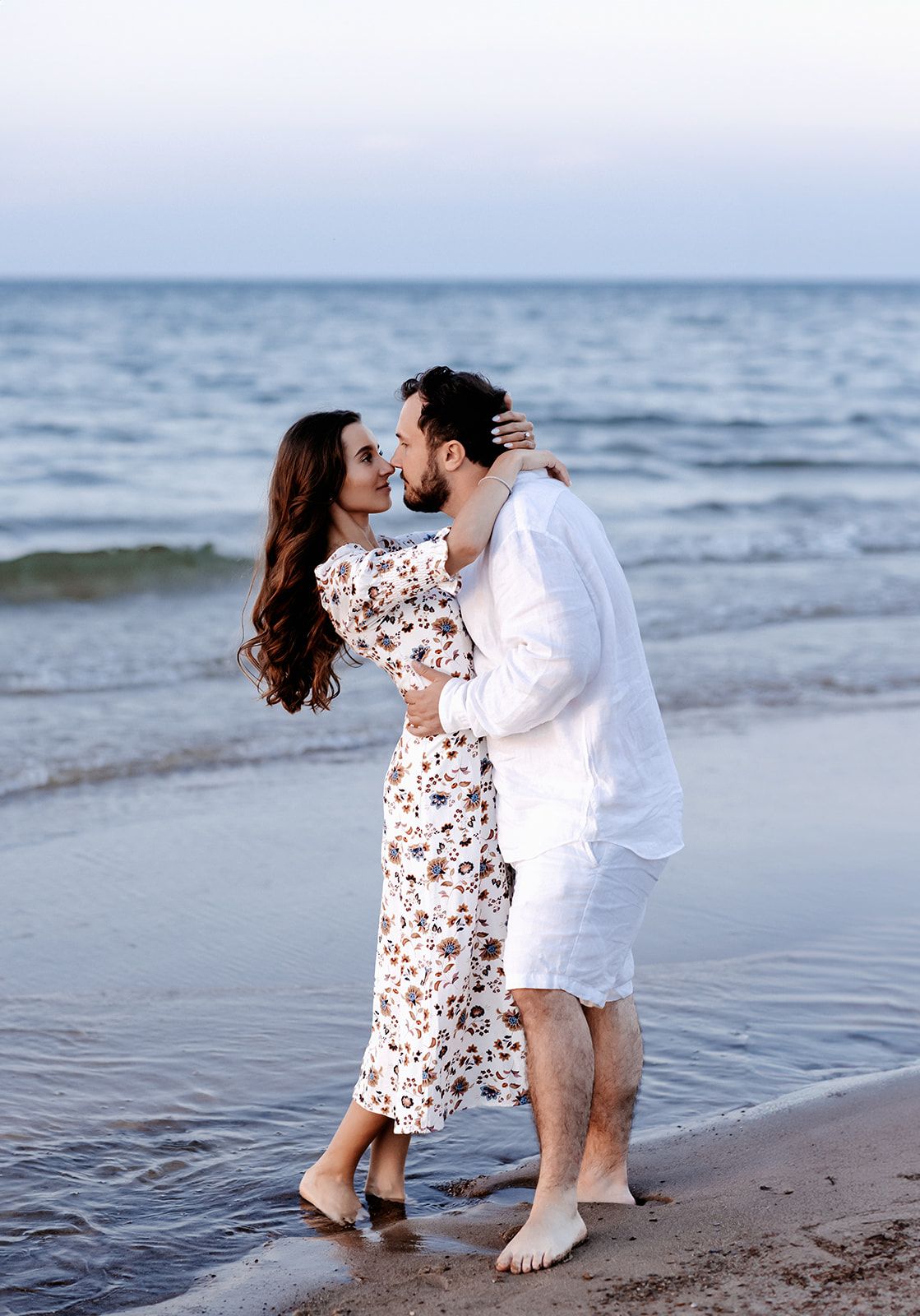 Romantic couple kissing barefoot on a beach shore with gentle ocean waves in the background at dusk.