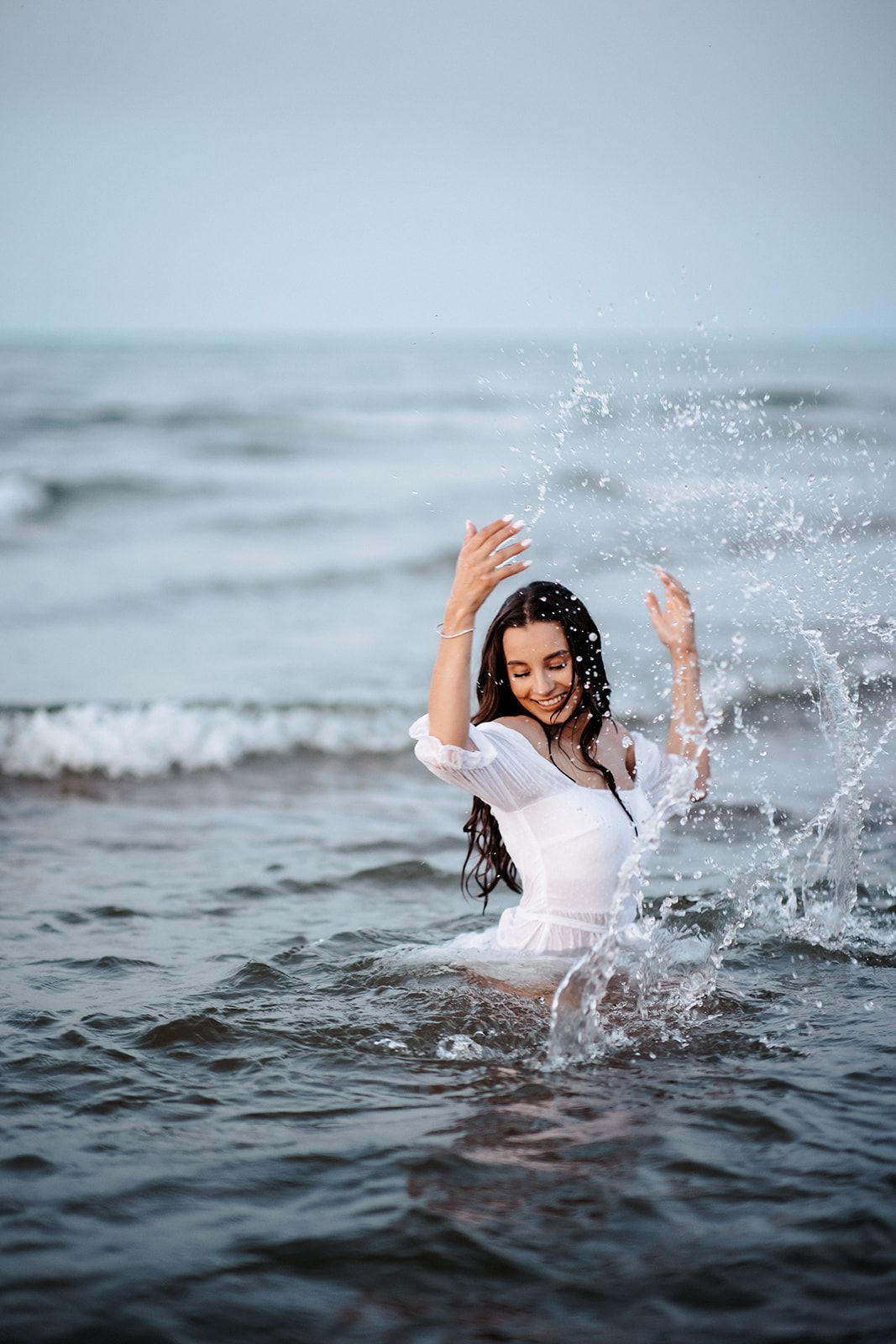 A joyful woman in a white dress splashing in ocean waves with her arms raised, smiling.
