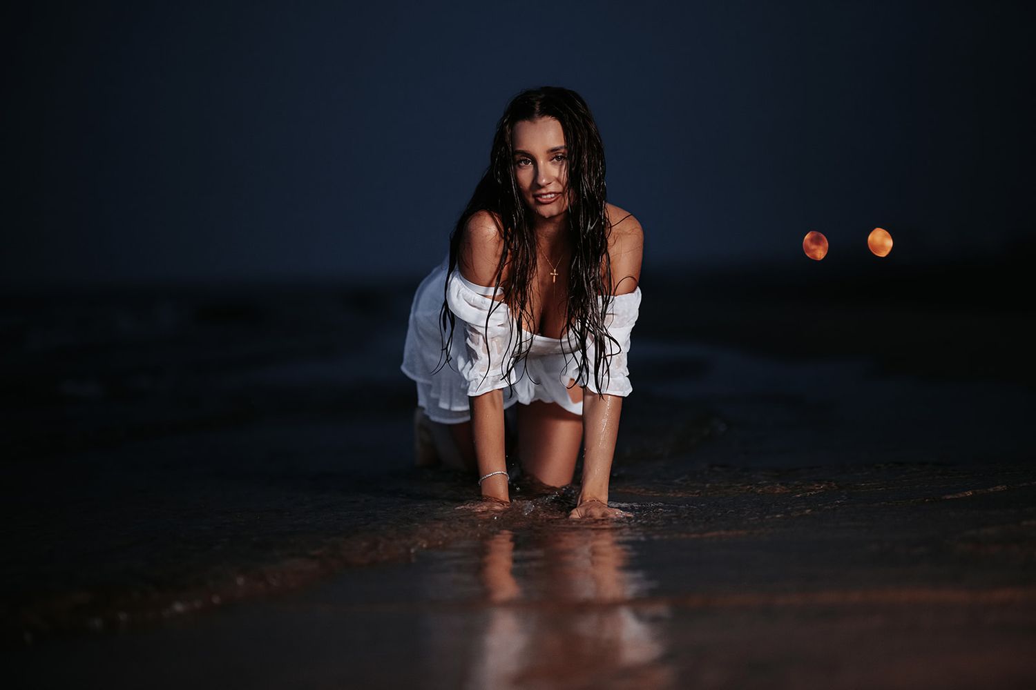 A woman in a white off-shoulder shirt kneels on a wet beach at night with bokeh lights in the background.