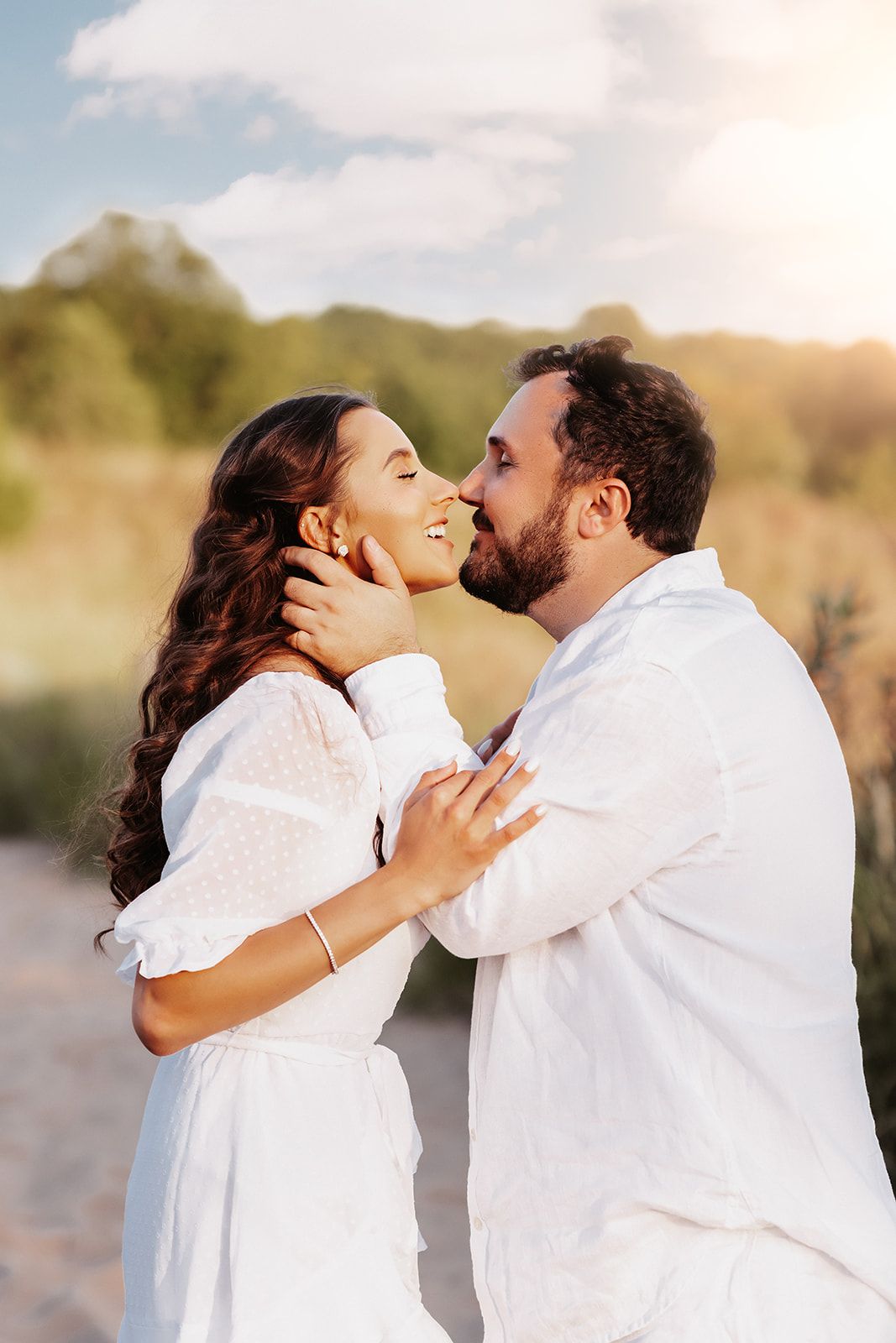 A bearded man in white gently holds a woman's face as they share a romantic kiss in golden sunlight.