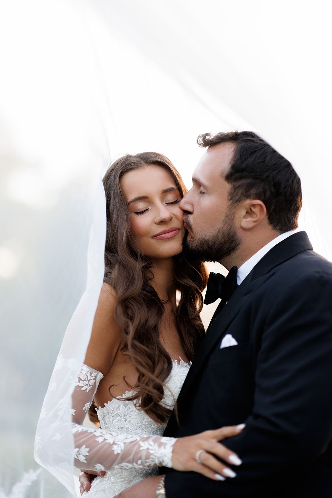 A groom in a black tuxedo kisses his bride on the cheek as she smiles with closed eyes beneath a flowing veil.