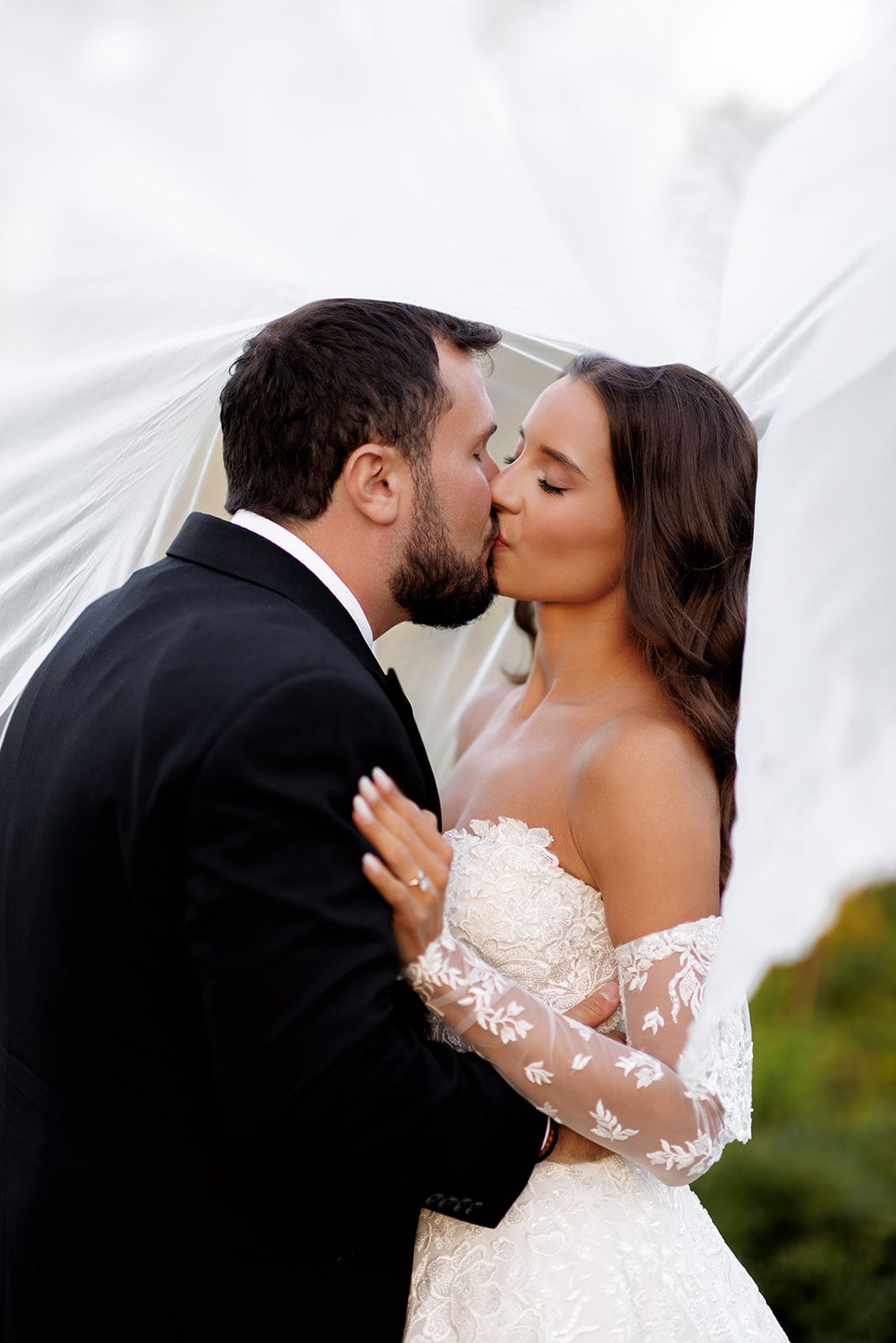 Bride and groom sharing a romantic kiss under a flowing white veil on their wedding day.