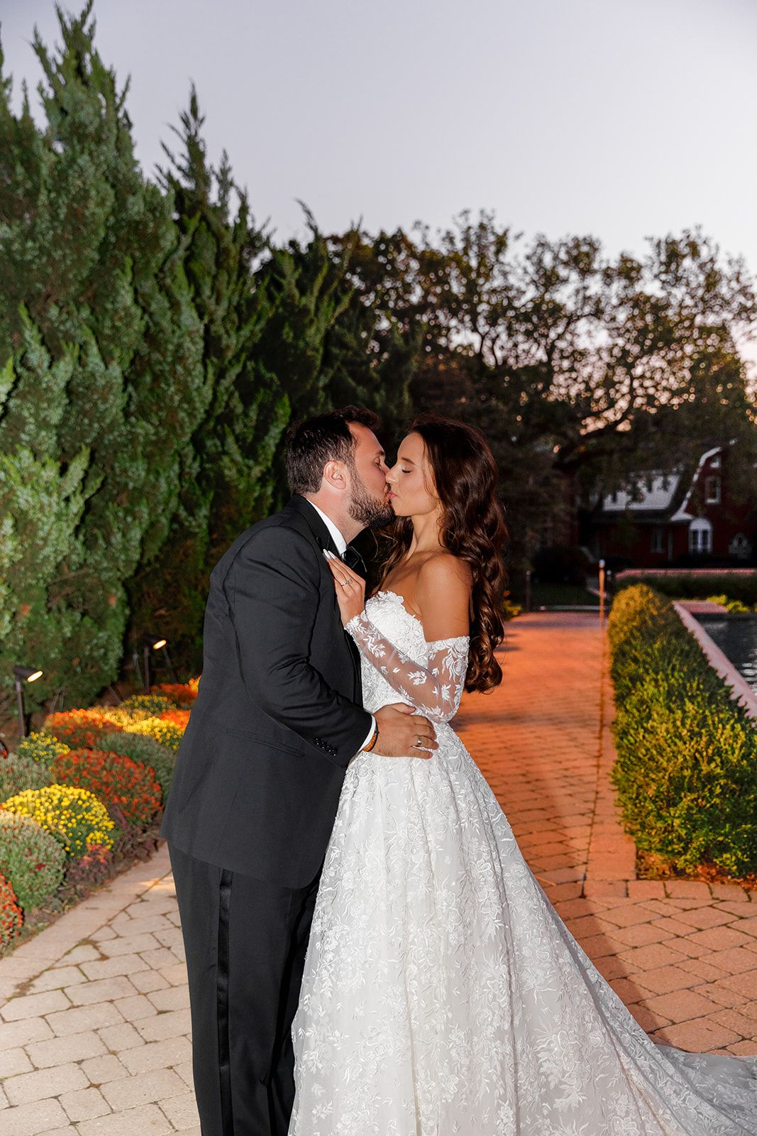 Newlyweds share a tender kiss at twilight in a beautifully landscaped garden, bride in stunning lace ball gown.