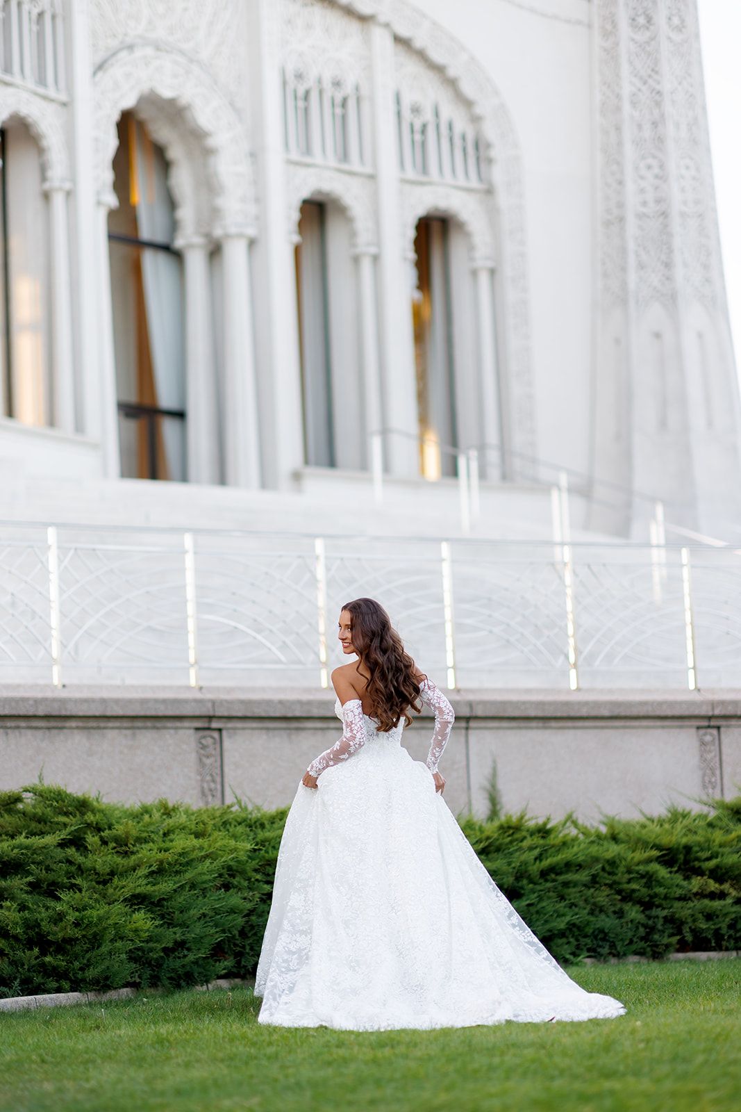 Bride in off-shoulder lace wedding gown with long sleeves stands on green lawn before ornate white building.