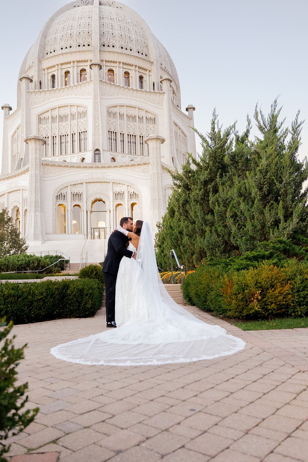 Bride and groom kissing in front of the Bahá'í House of Worship, bride wearing a long cathedral veil.