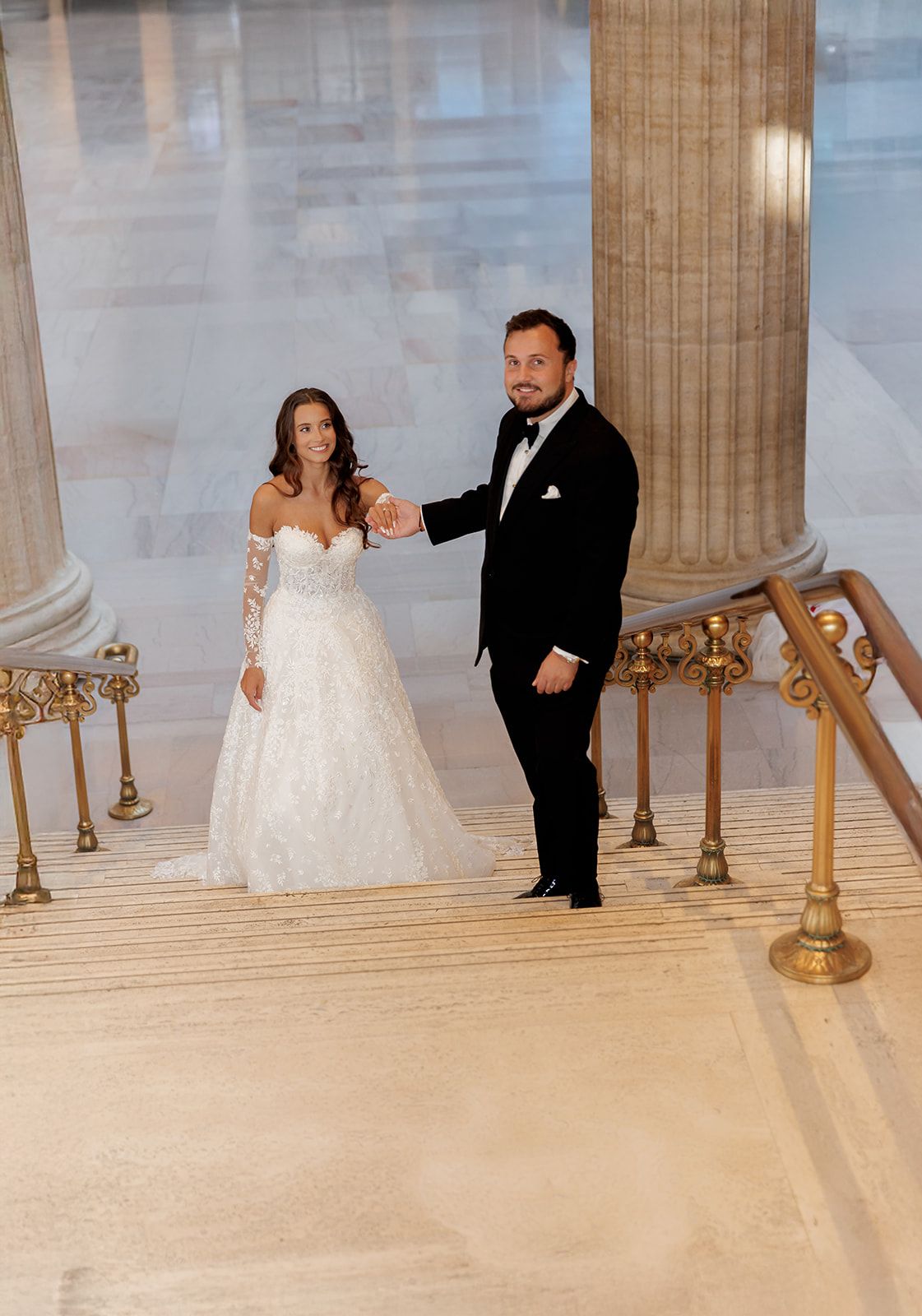 Bride in lace ball gown and groom in tuxedo pose on grand marble staircase with gold railings.