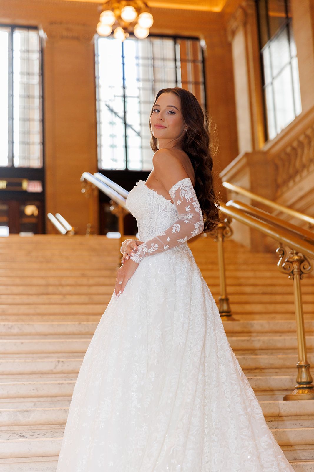 Bride in off-shoulder lace wedding gown on grand staircase with gold railings.