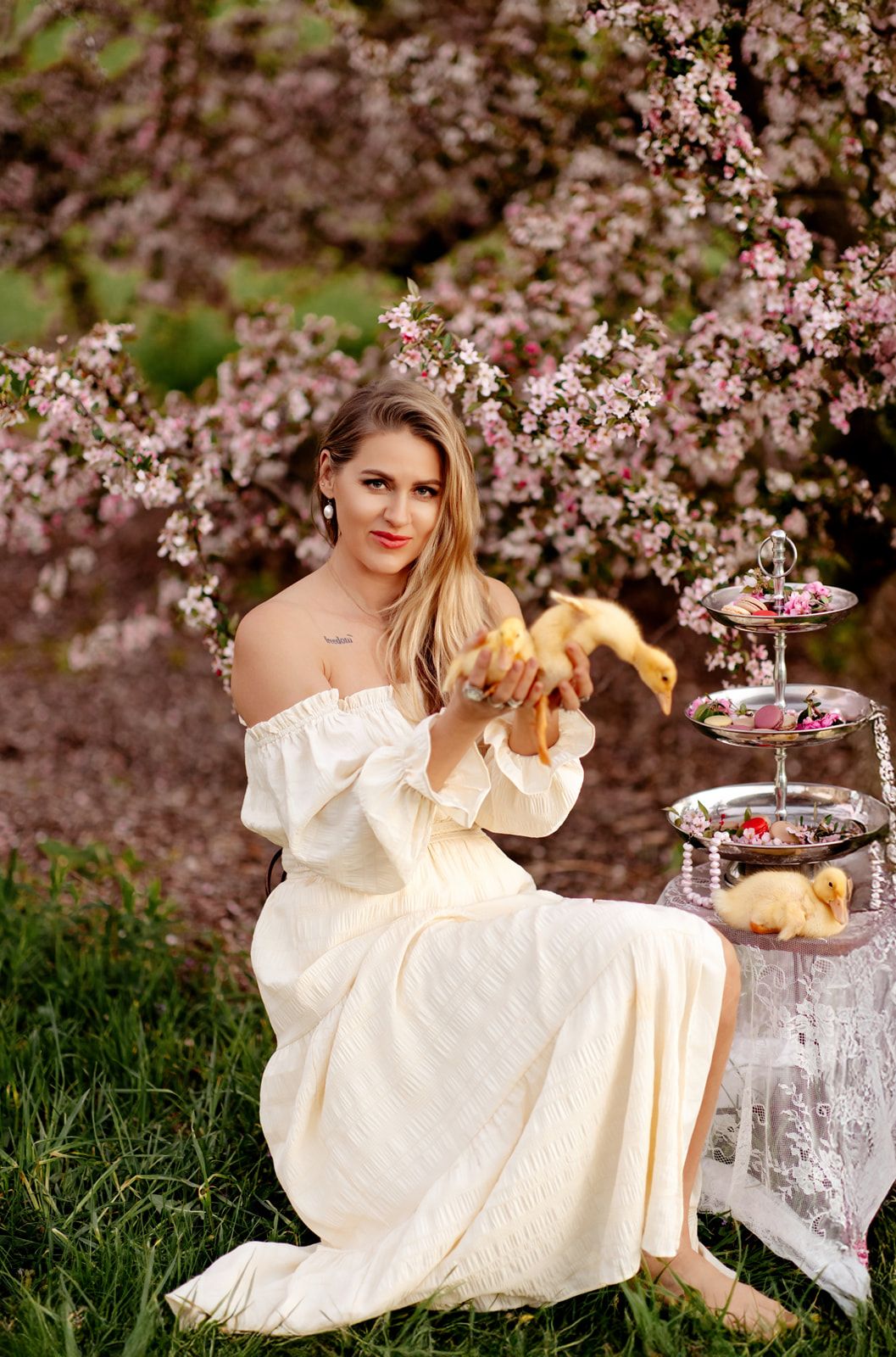 Woman in cream off-shoulder dress holds ducklings near tiered tray under pink cherry blossom tree.