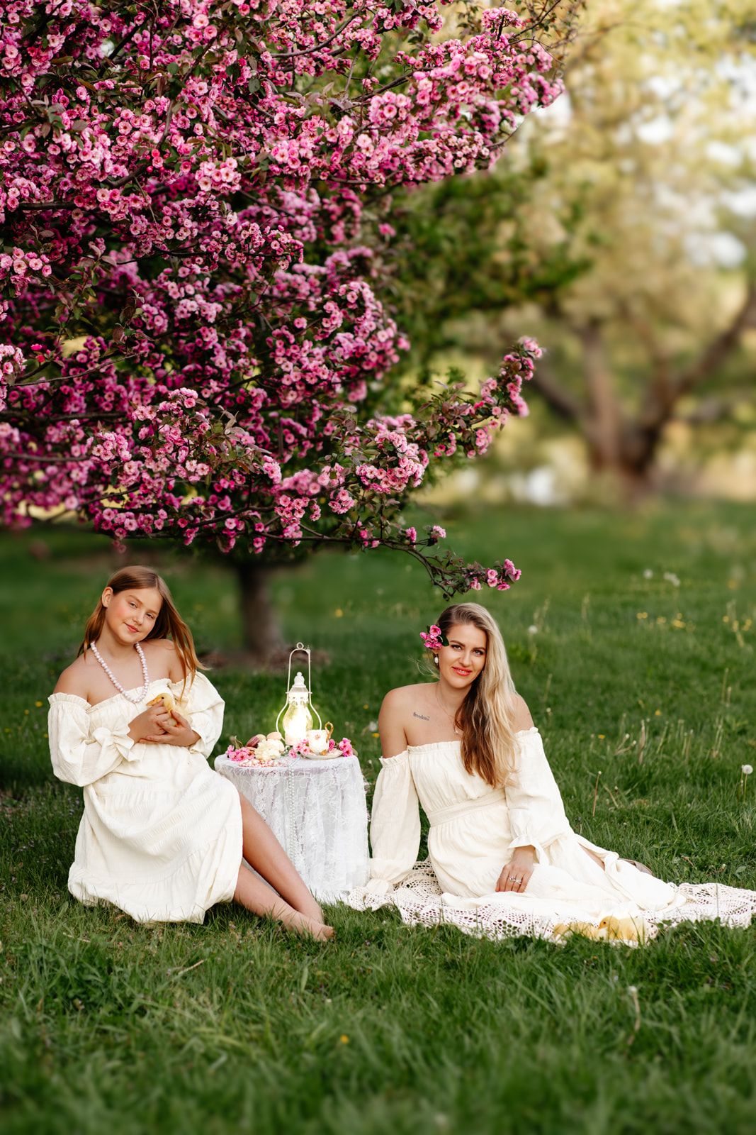 Two women in white dresses enjoy a picnic under pink blossoming trees on green grass.
