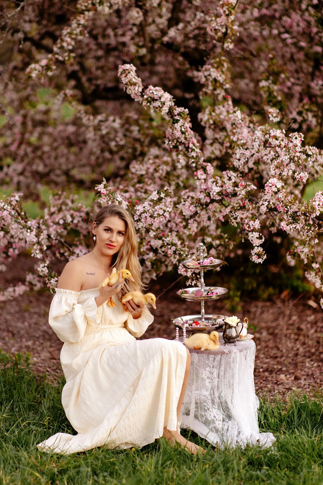 Woman in white off-shoulder dress sits by blooming cherry tree with tiered dessert stand on tree stump.