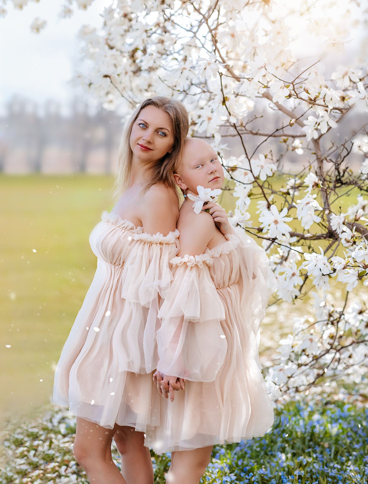 Mother and baby in matching blush tulle dresses pose by blooming white magnolia tree outdoors in spring.