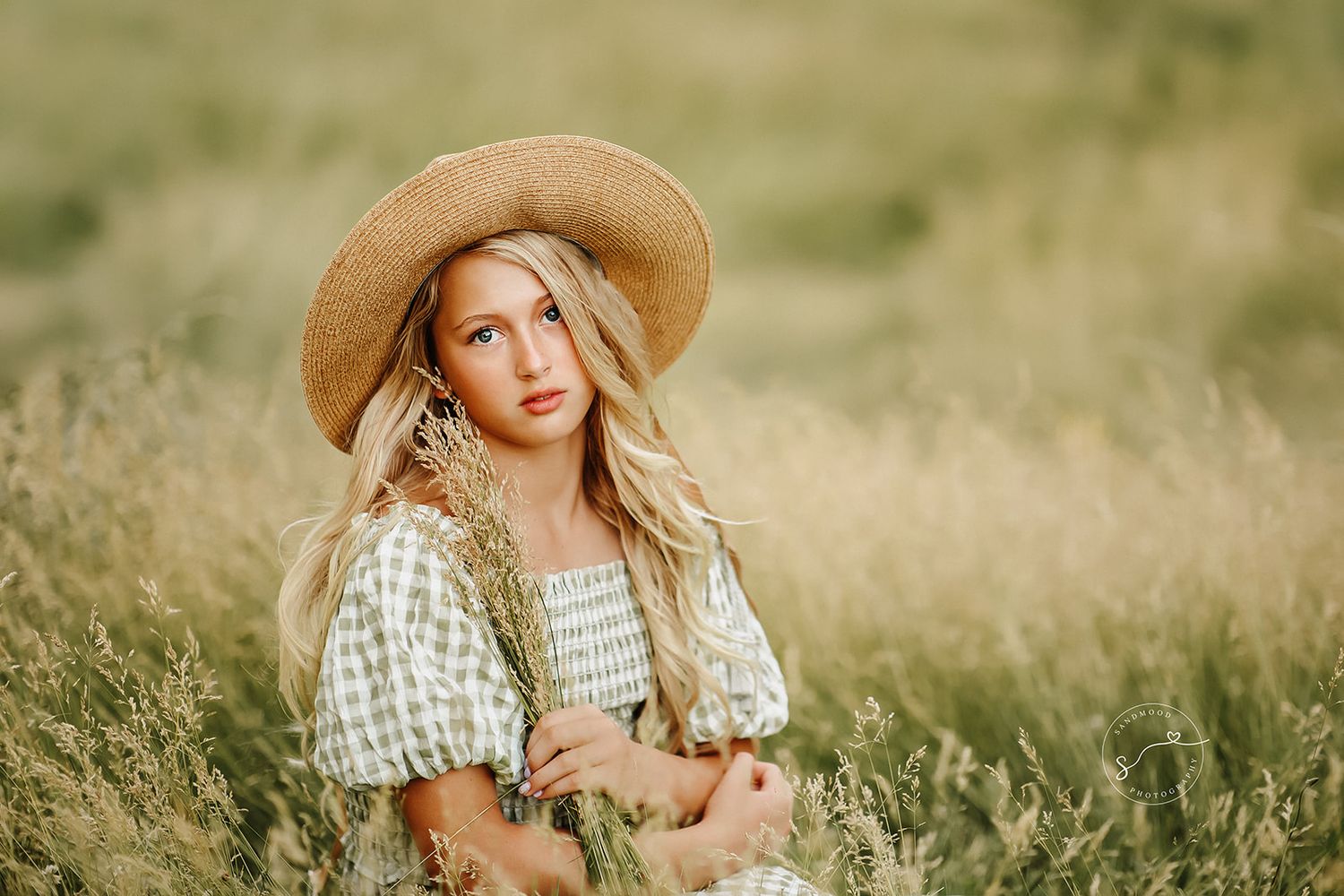 Young blonde woman in straw hat holding wheat bouquet while sitting in golden meadow field.