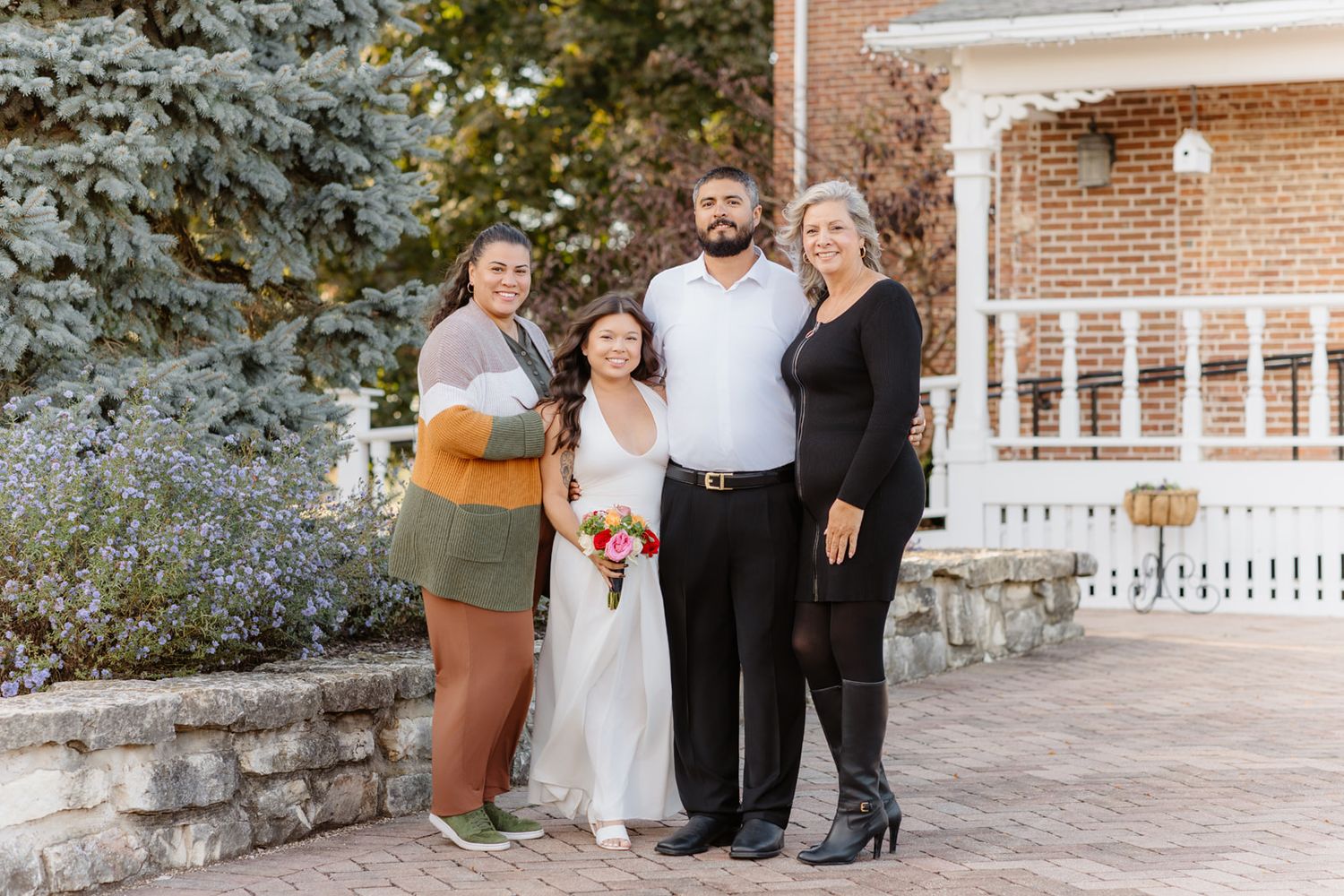 Bride and groom pose with two female family members on brick patio near blue spruce tree at wedding.