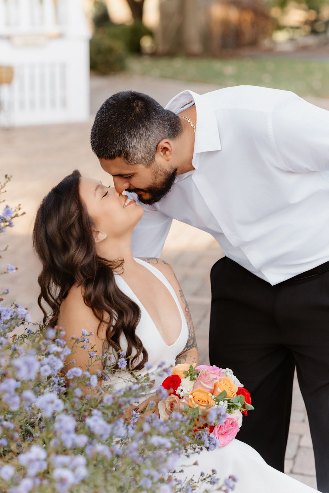 Bride and groom sharing romantic kiss outdoors near purple wildflowers, bride holding vibrant multicolored rose bouquet.