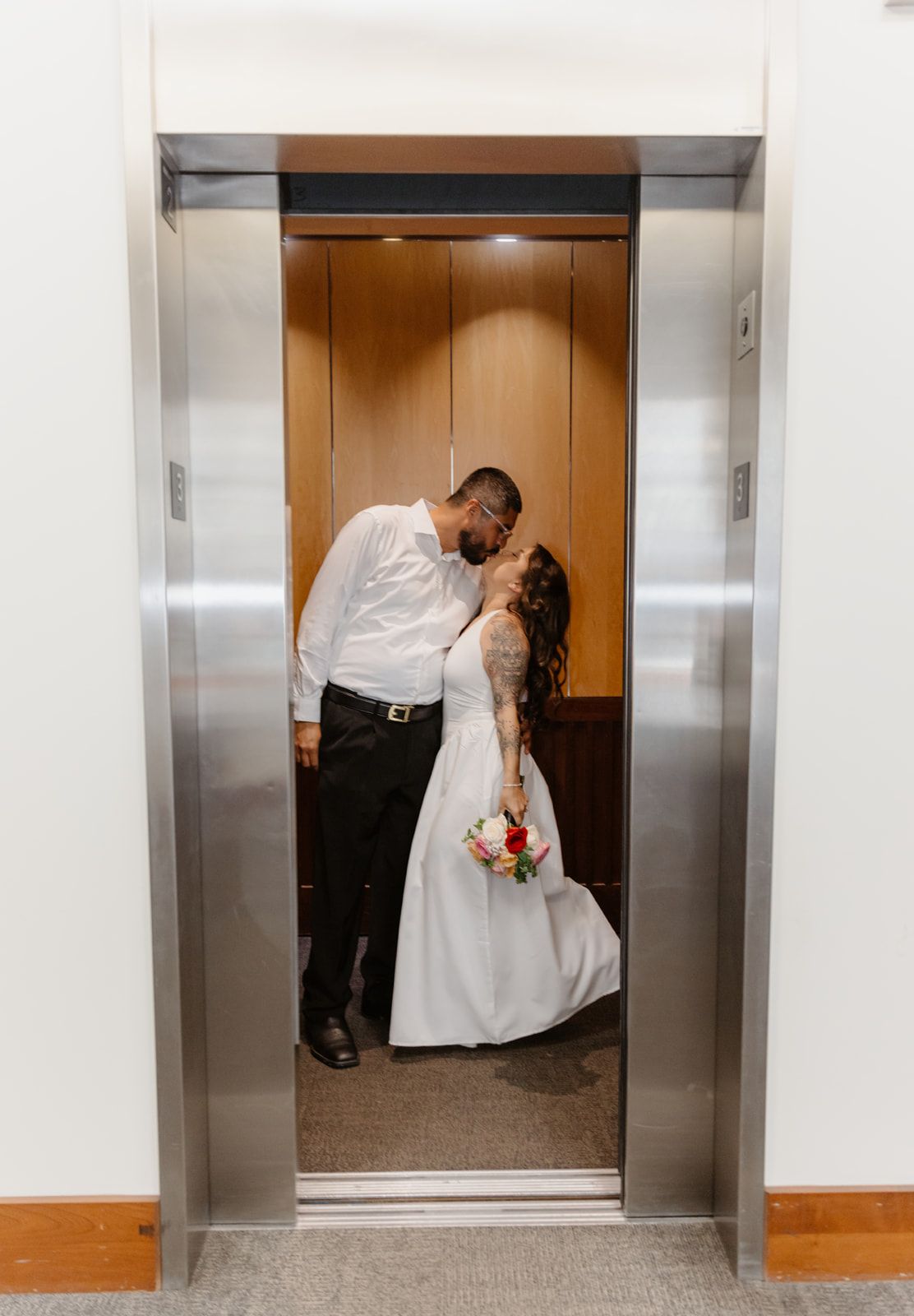 Bride and groom sharing a romantic kiss inside an elevator, bride holding a colorful bouquet in a white wedding dress.