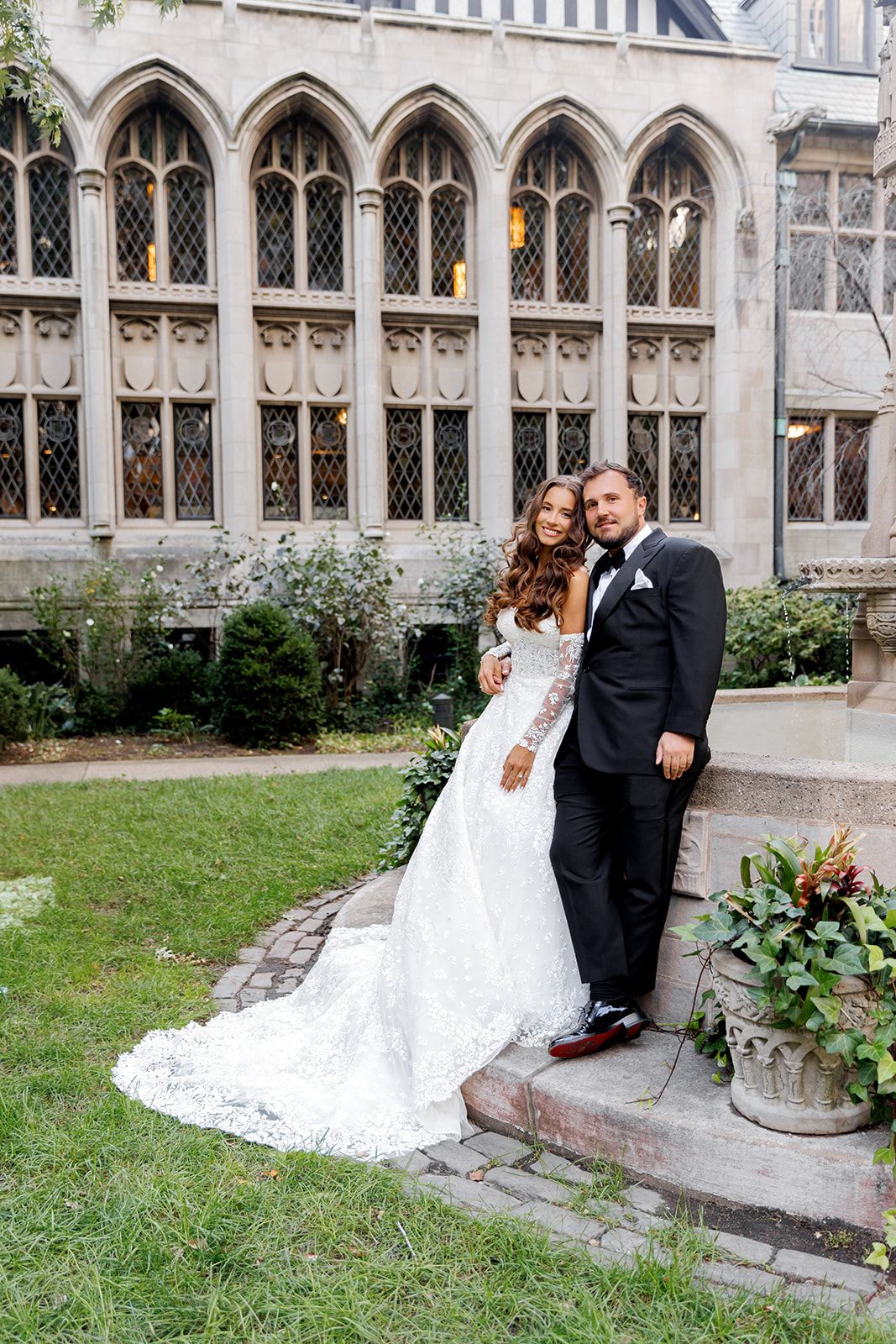 Wedding couple embracing in front of Gothic arched windows, bride wearing elegant lace train gown.