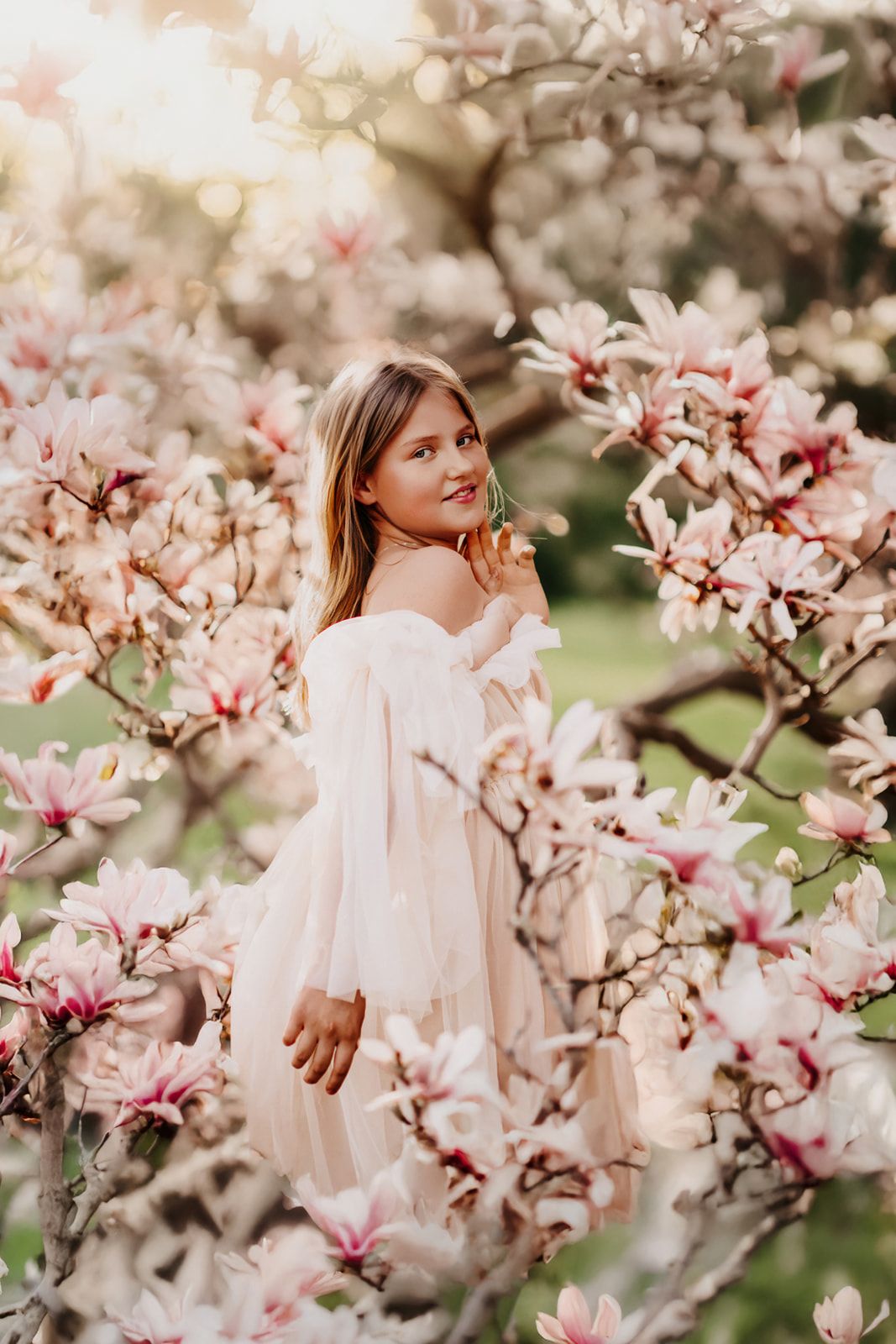 Young girl in white dress surrounded by pink magnolia blossoms in golden hour light.