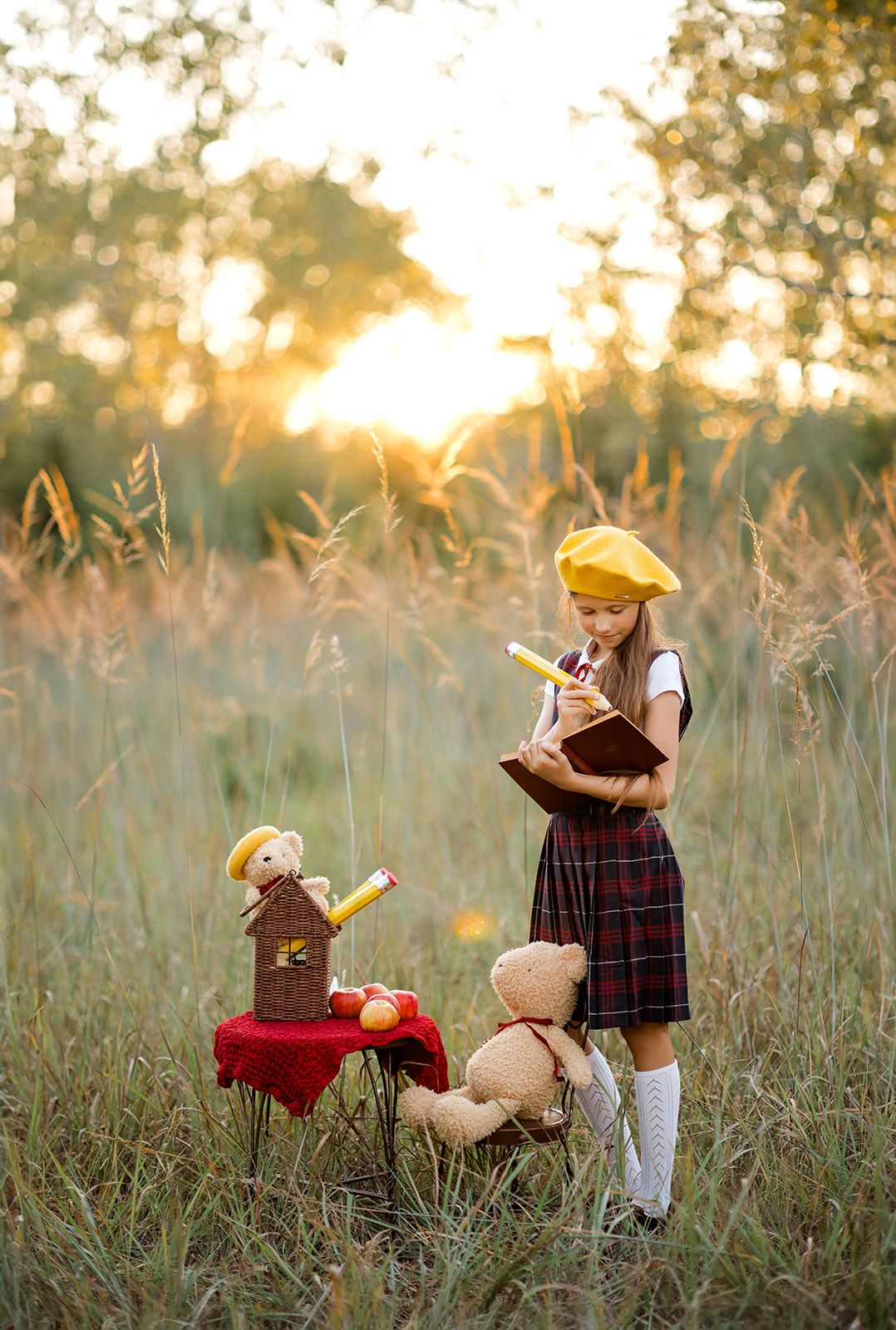 Girl in yellow beret reads book outdoors at sunset with teddy bears and small table in golden field.