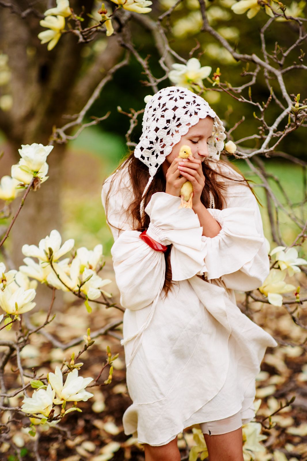 Sweet girl in white linen dress and lace bonnet holds a yellow chick near magnolia blossoms.