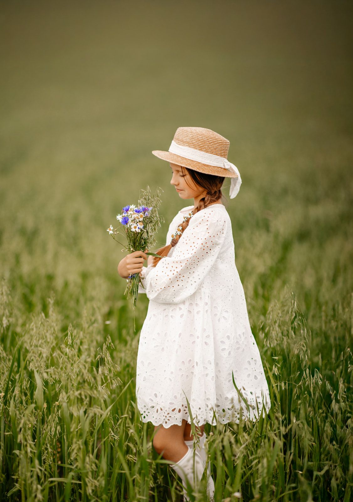 Young girl in white lace dress and straw hat holding wildflower bouquet in a lush green field.
