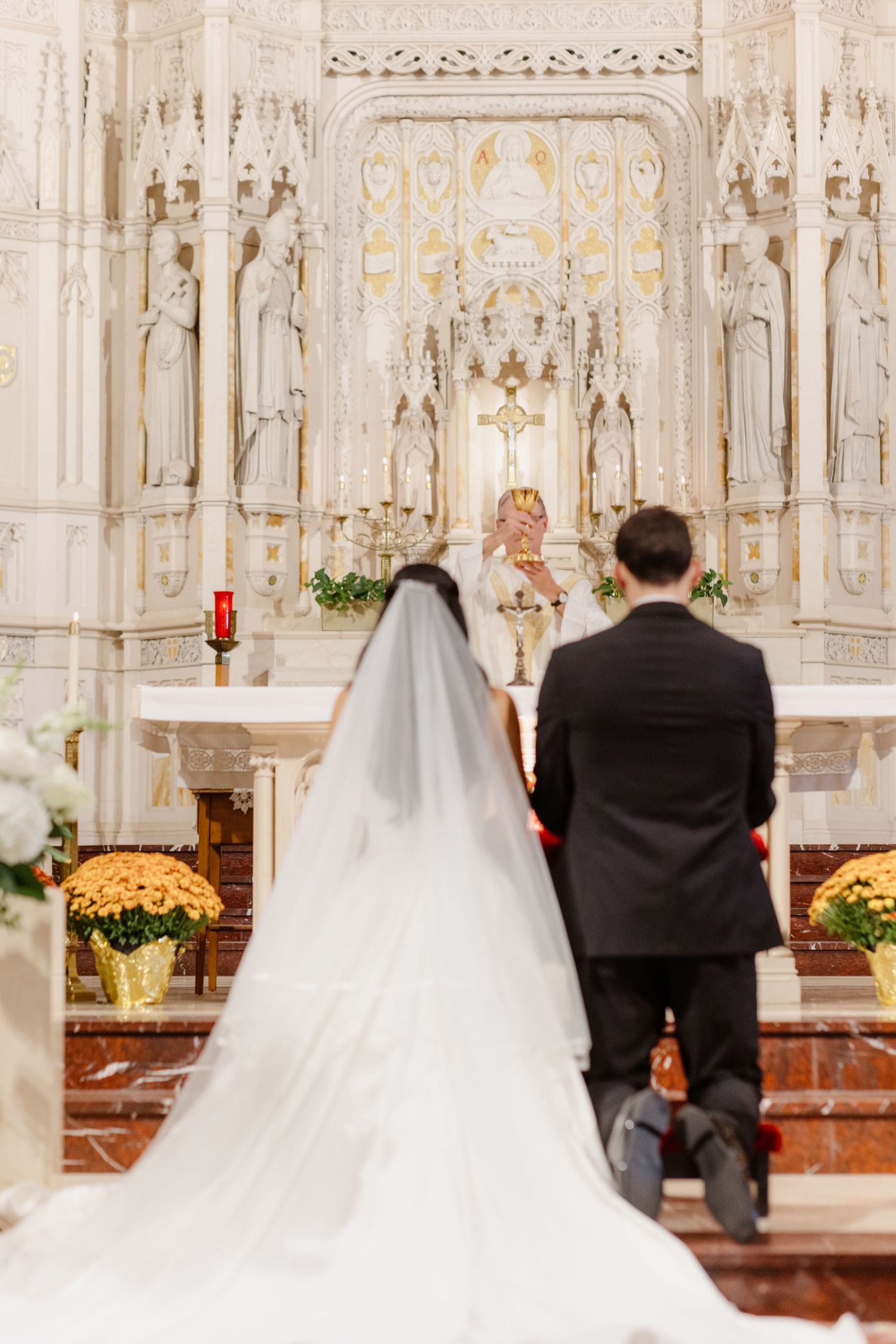 Bride and groom kneel before ornate white altar during traditional church wedding ceremony.