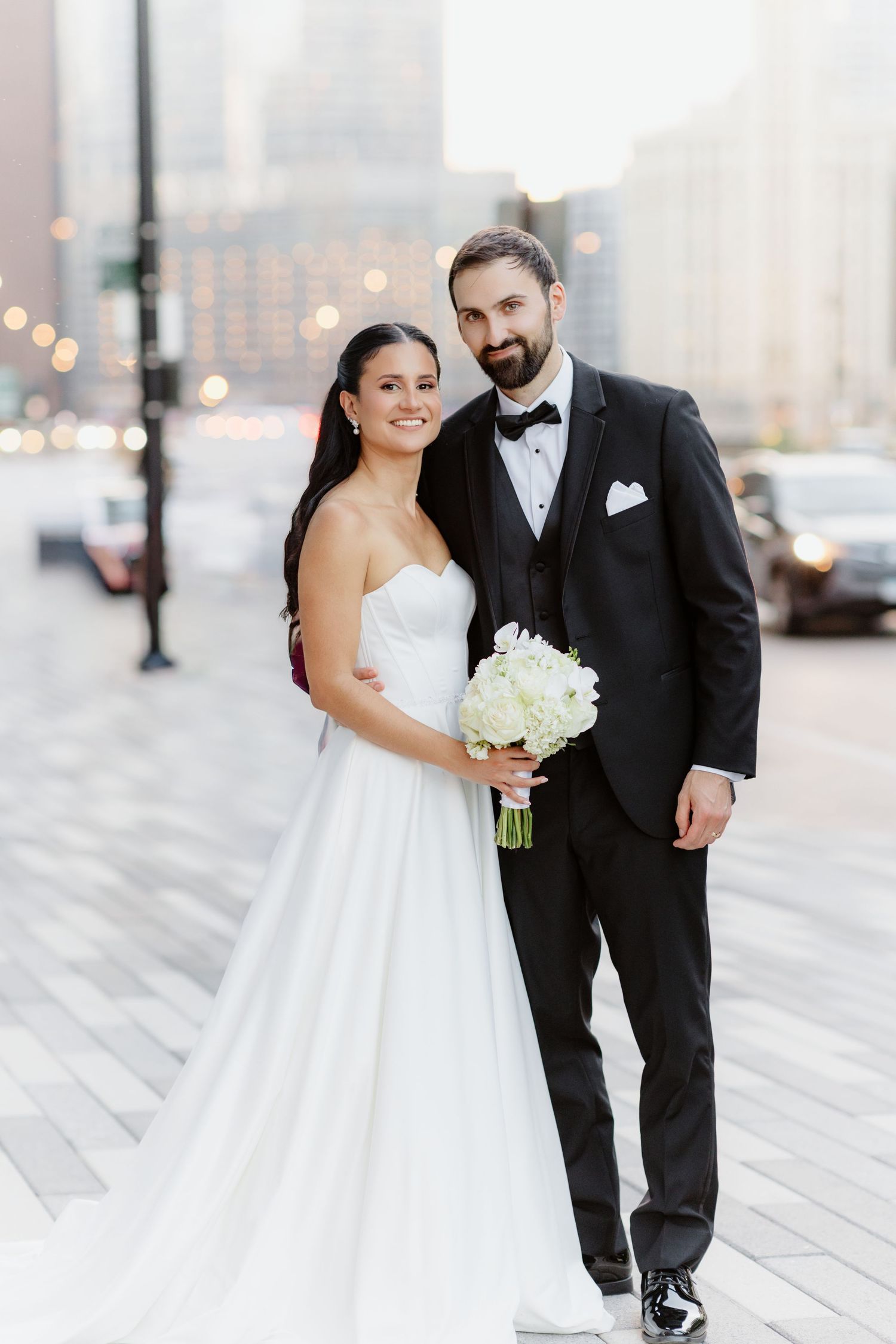 Bride in strapless white gown holds white bouquet beside groom in black tuxedo on urban street at dusk.