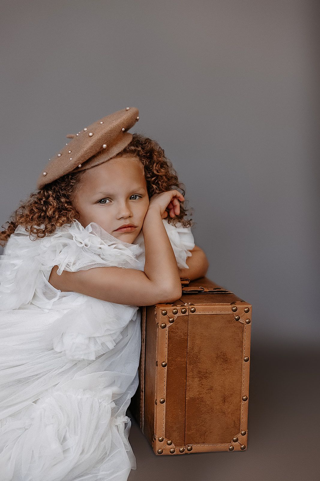 Young curly-haired girl in white dress and beige beret leaning on vintage brown suitcase.