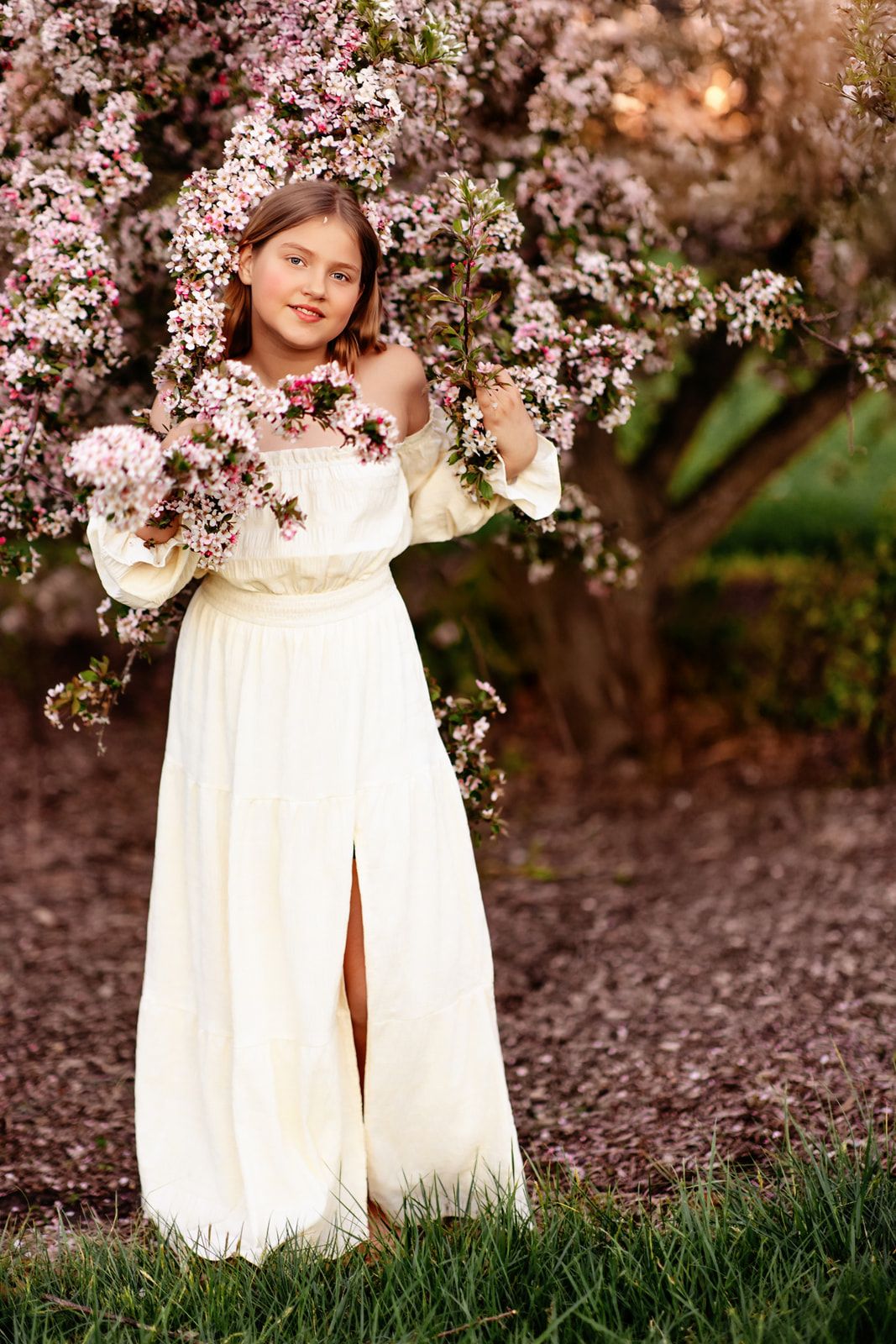 Young girl in elegant white off-shoulder gown poses among pink blossoming spring tree branches.