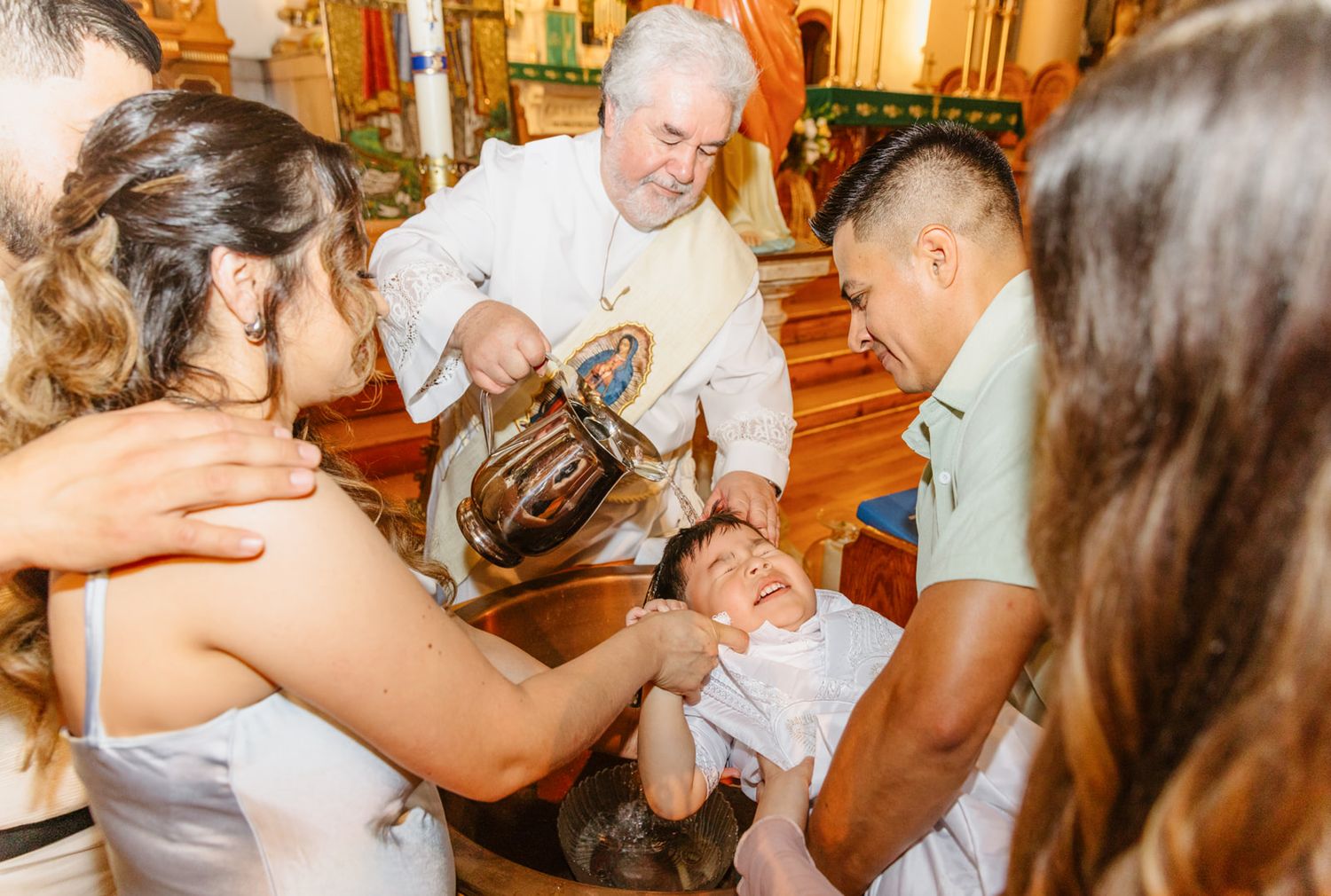 Priest performing Catholic baptism ceremony, pouring holy water over infant held by father in ornate church.